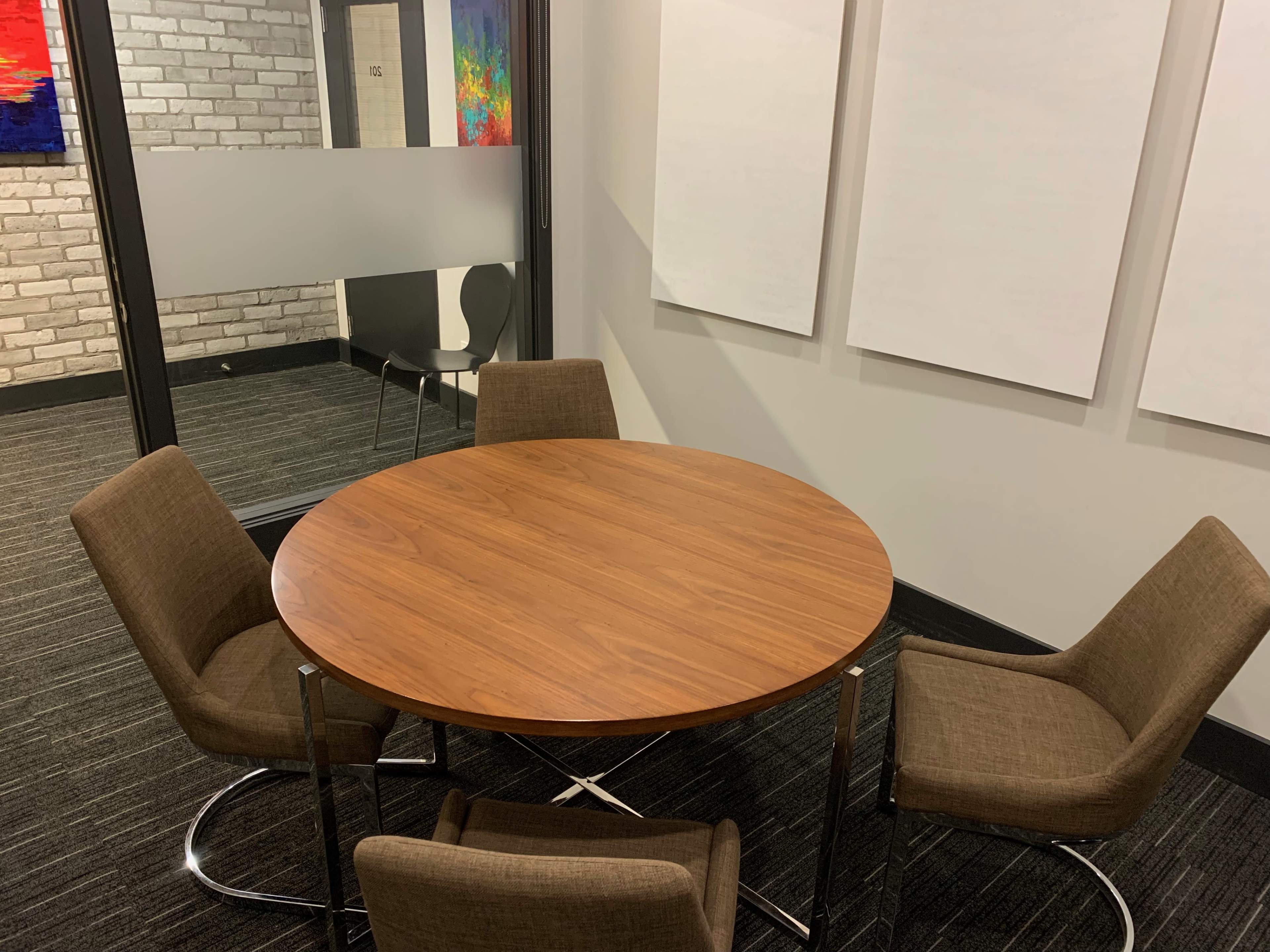 A round wooden table surrounded by four brown chairs in a modern office space with a brick wall and whiteboard surfaces.