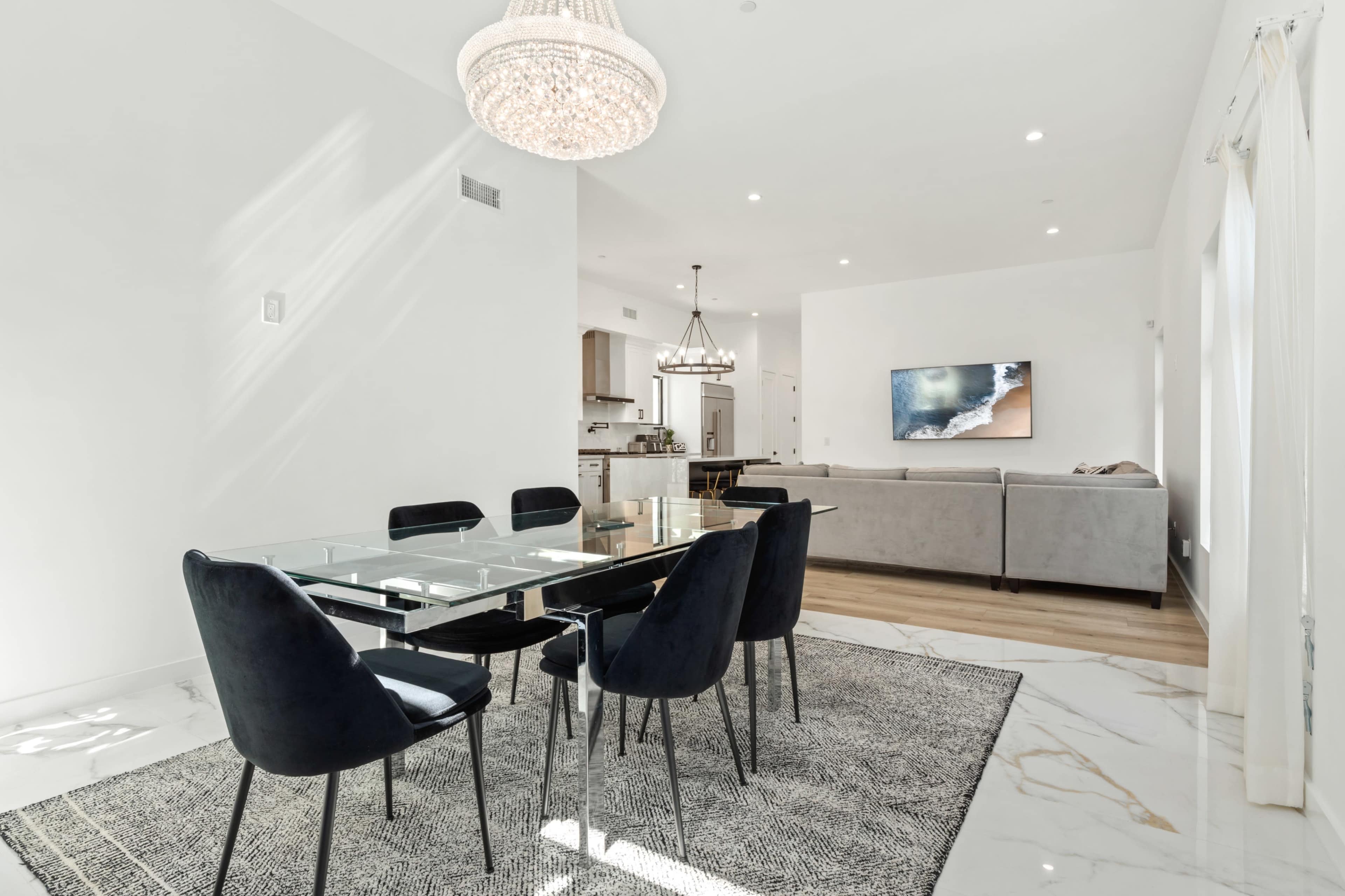 The image shows a modern dining area with a glass table surrounded by black chairs, adjacent to a living room featuring a gray sofa and a large wall-mounted television.