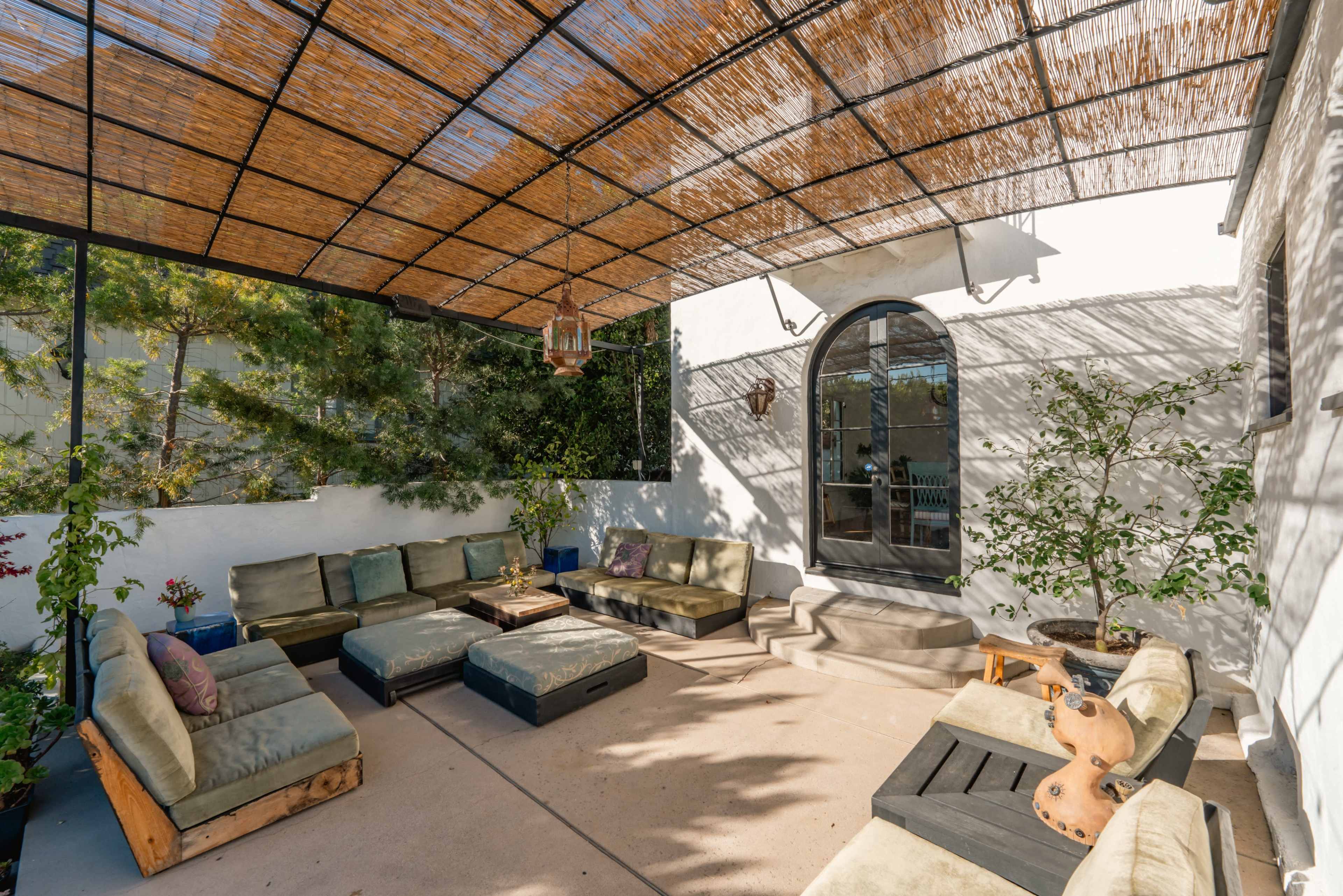 The image shows a spacious outdoor seating area with cushioned benches arranged around a wooden table, shaded by a bamboo canopy and bordered by plants.