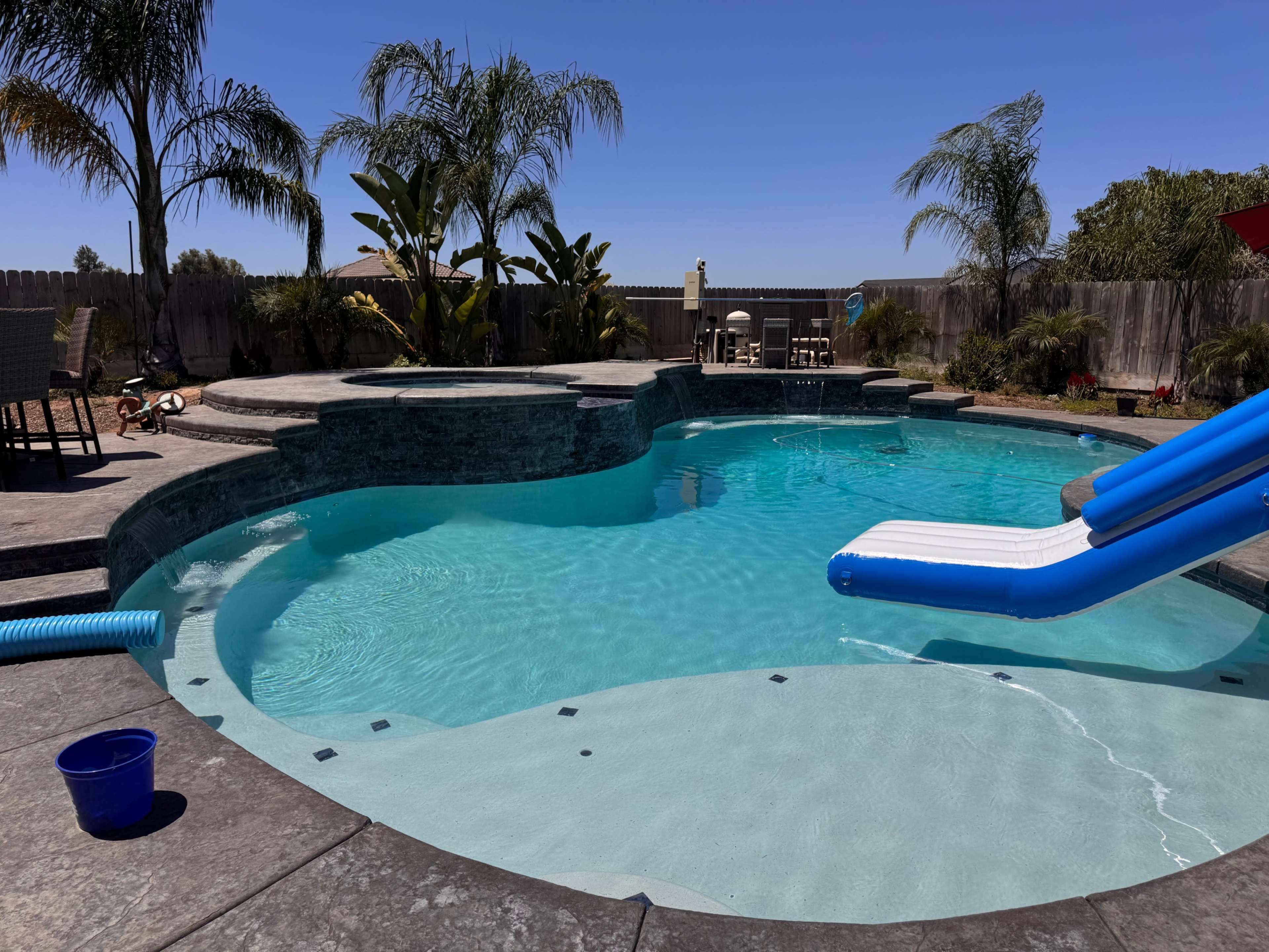 The image shows a backyard pool area with a water slide, surrounded by palm trees and lounge chairs.
