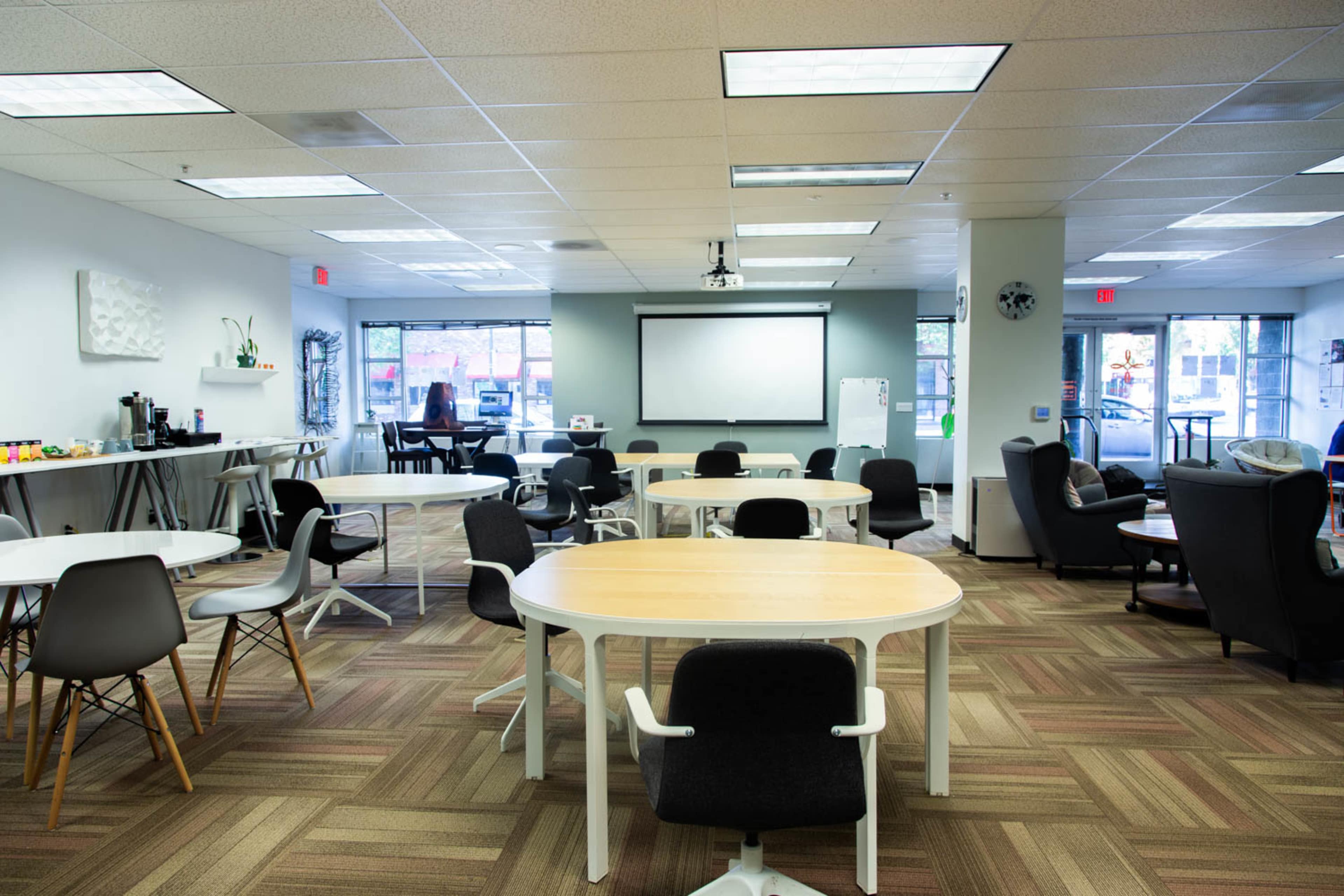 A modern office space with several round and rectangular tables, chairs, and a presentation screen at the front.