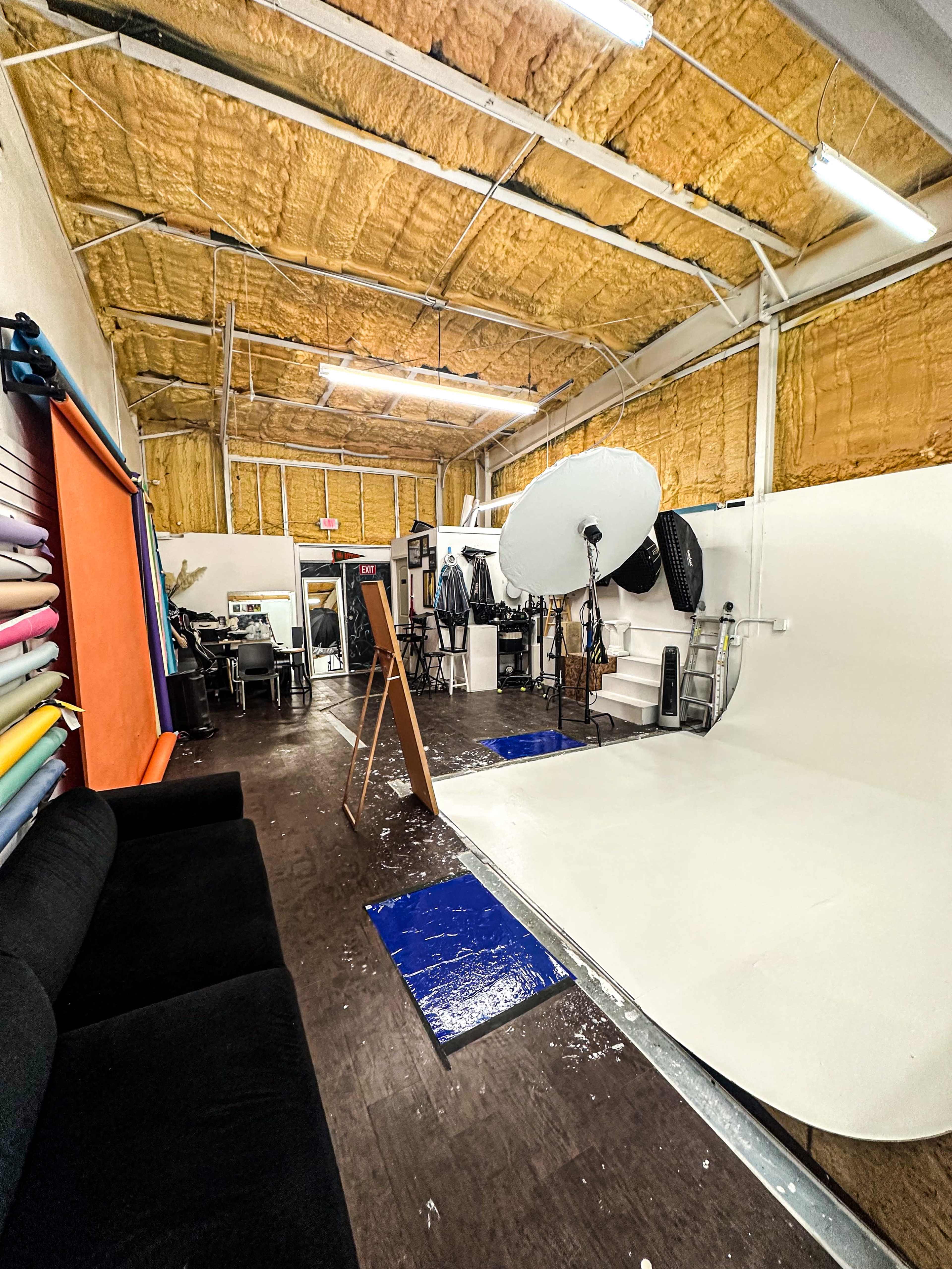 The image shows a photo studio with a white backdrop, various lighting equipment, and a seating area, all set within a space featuring insulated walls and a wooden floor.