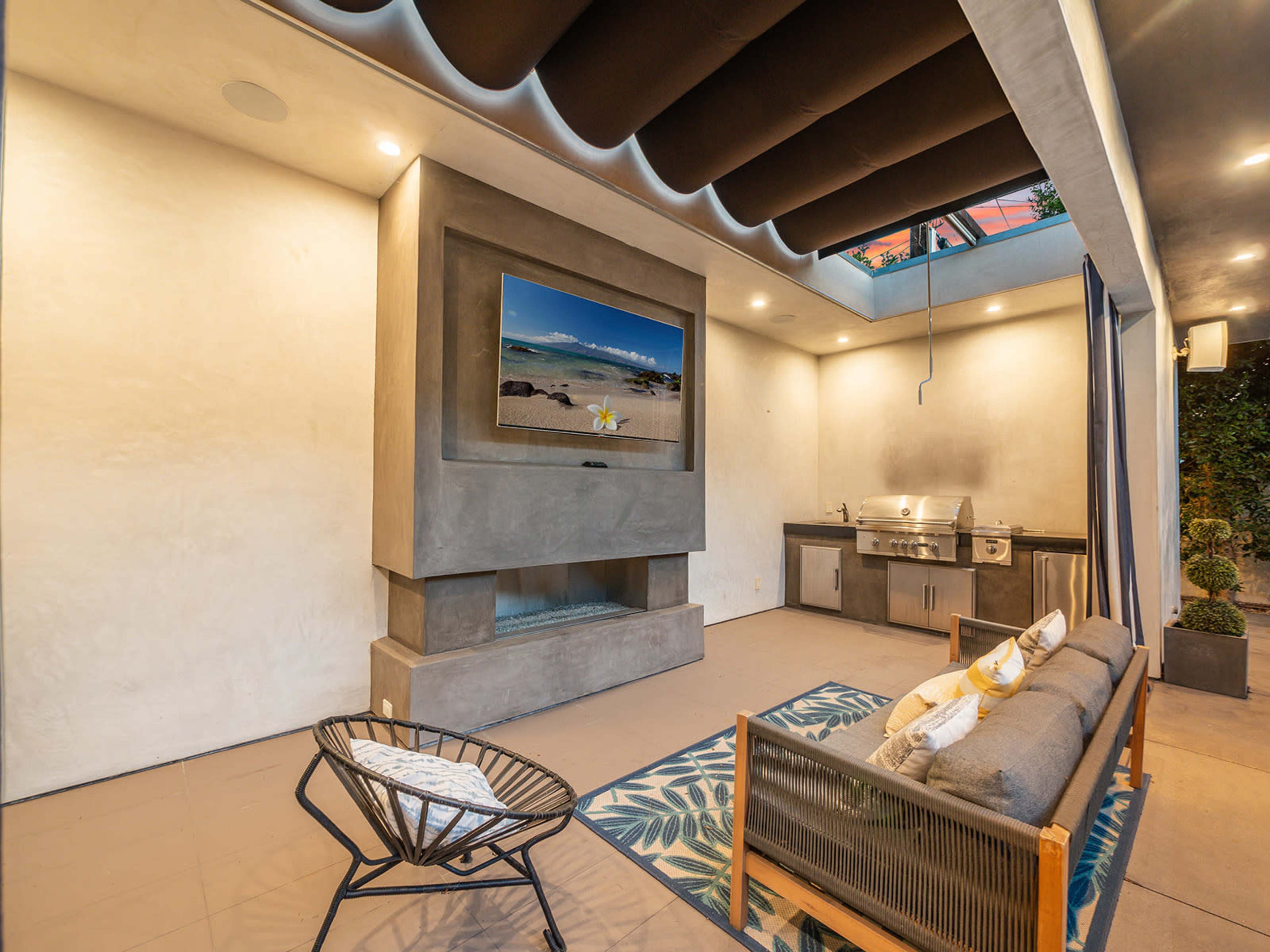 An outdoor living space with a sofa, a television mounted on a concrete wall, and an outdoor kitchen area.