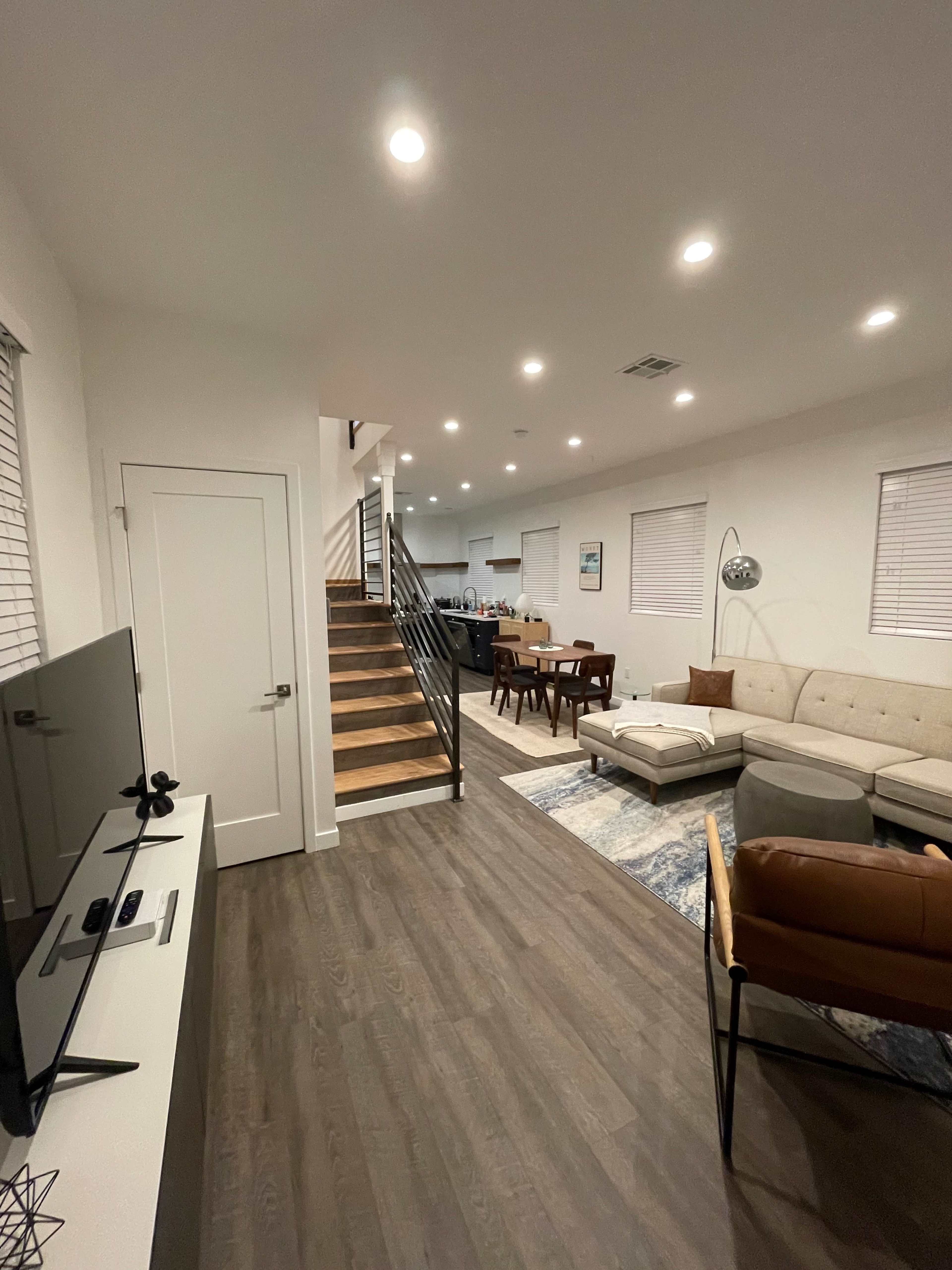 The image shows a modern living space with a staircase on the left, a couch and television in the foreground, and a dining area in the background.