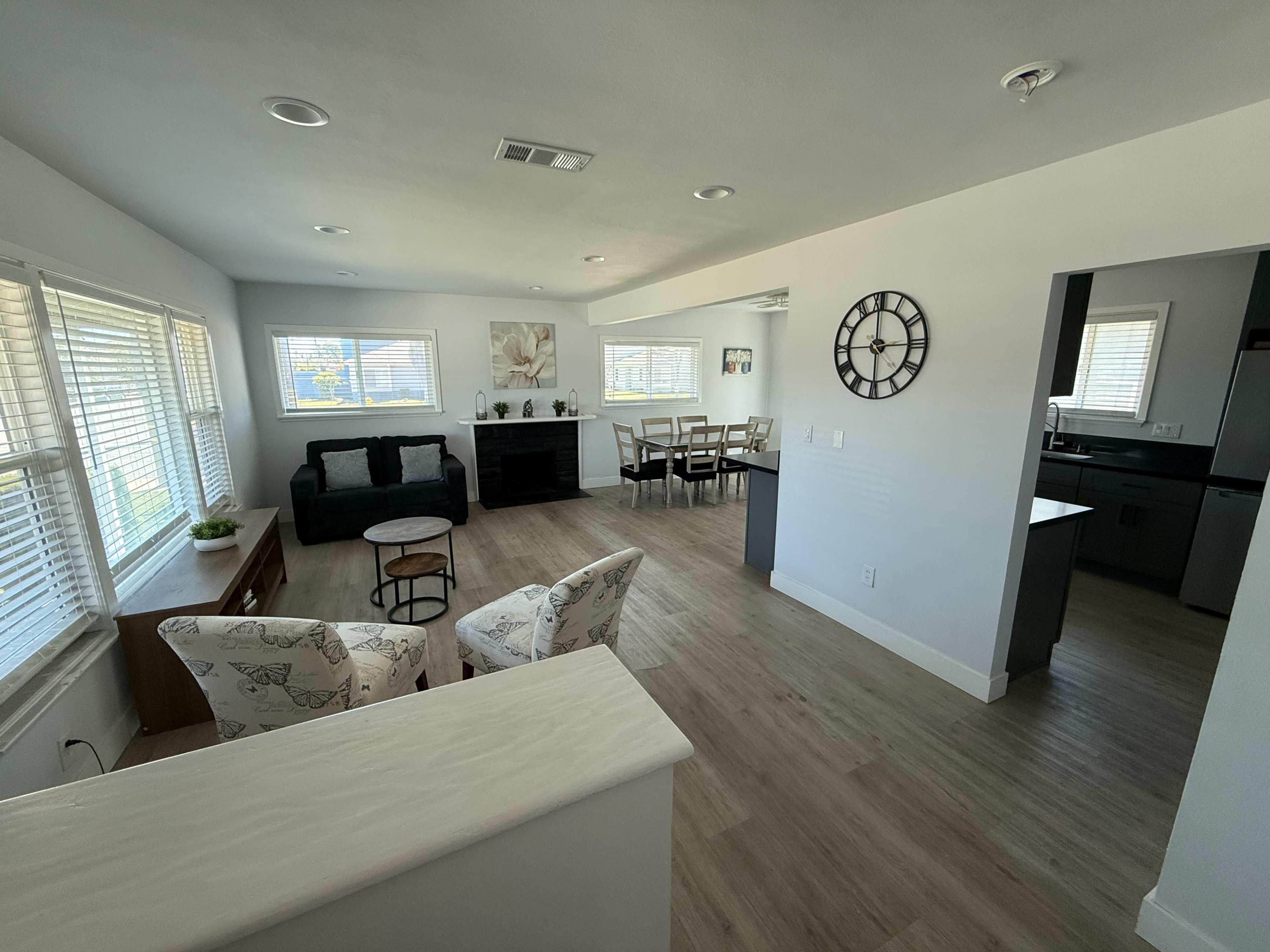 The image shows a bright, modern living space with a seating area, dining table, and kitchen visible in an open floor plan layout.