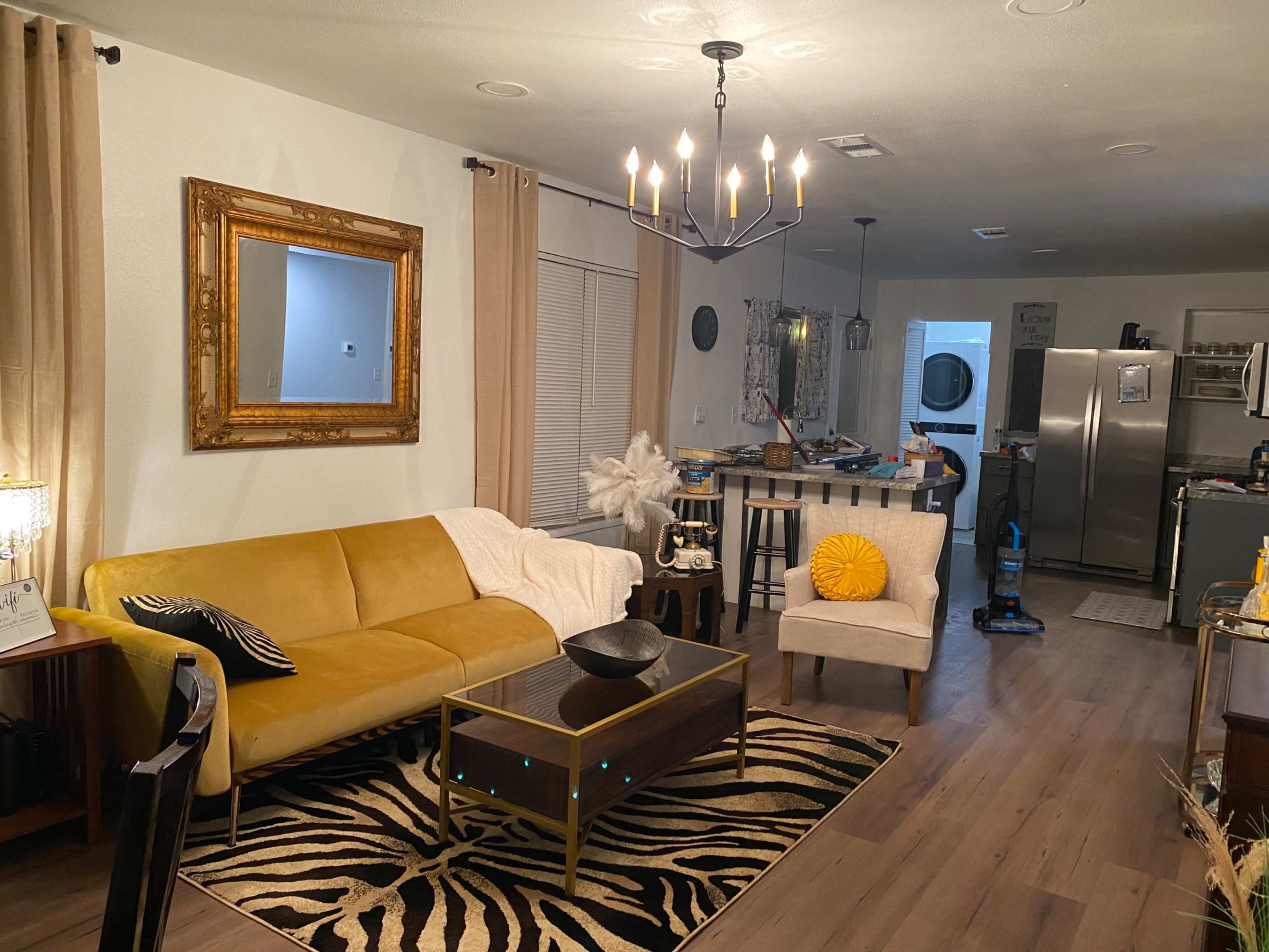 A living room featuring a yellow couch, a coffee table, a white chair with a decorative pillow, and a chandelier overhead, with a kitchen visible in the background.