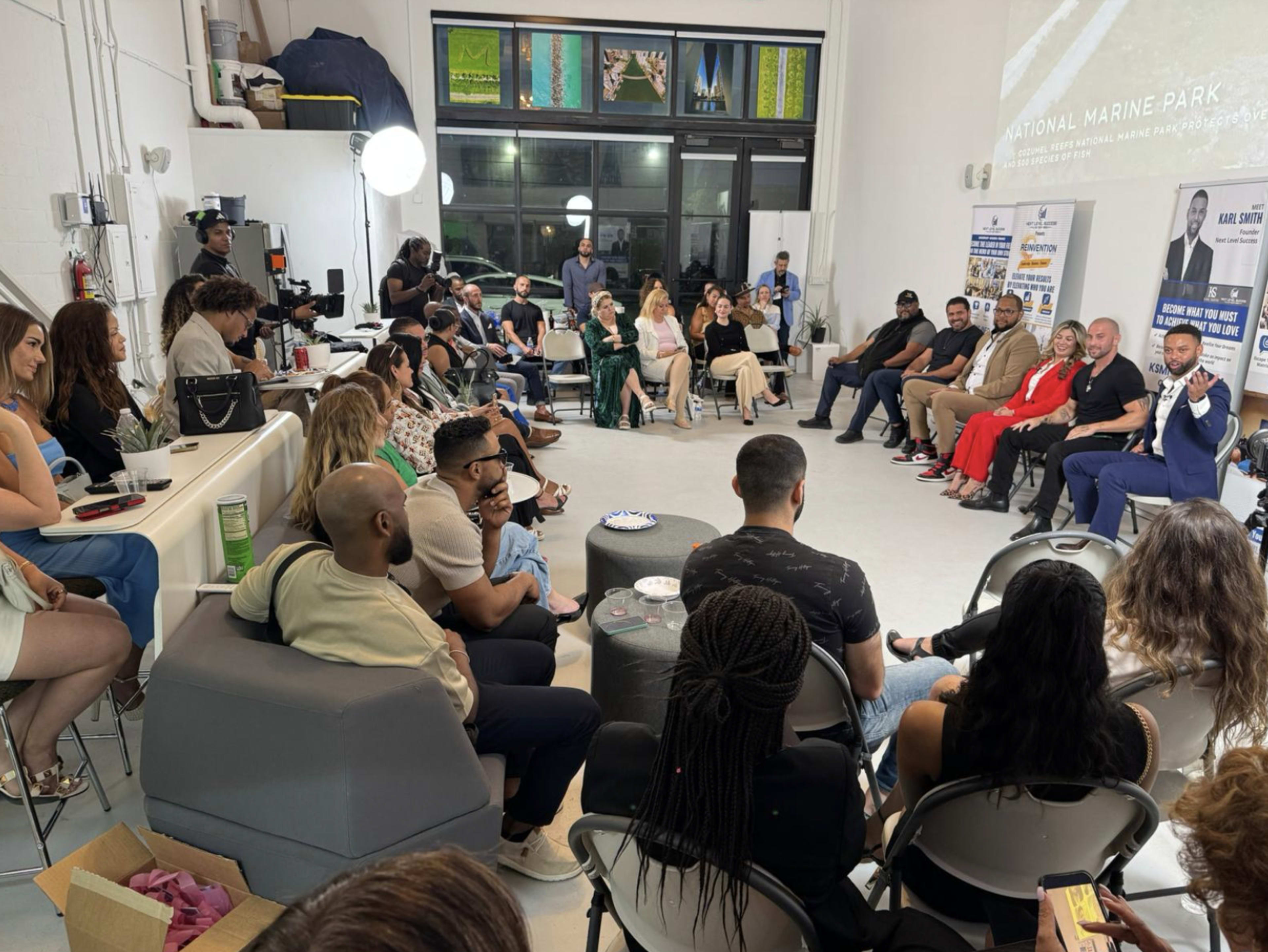 A diverse group of people is seated in a well-lit space, engaged in a discussion or presentation, with some individuals on stage and others in the audience.