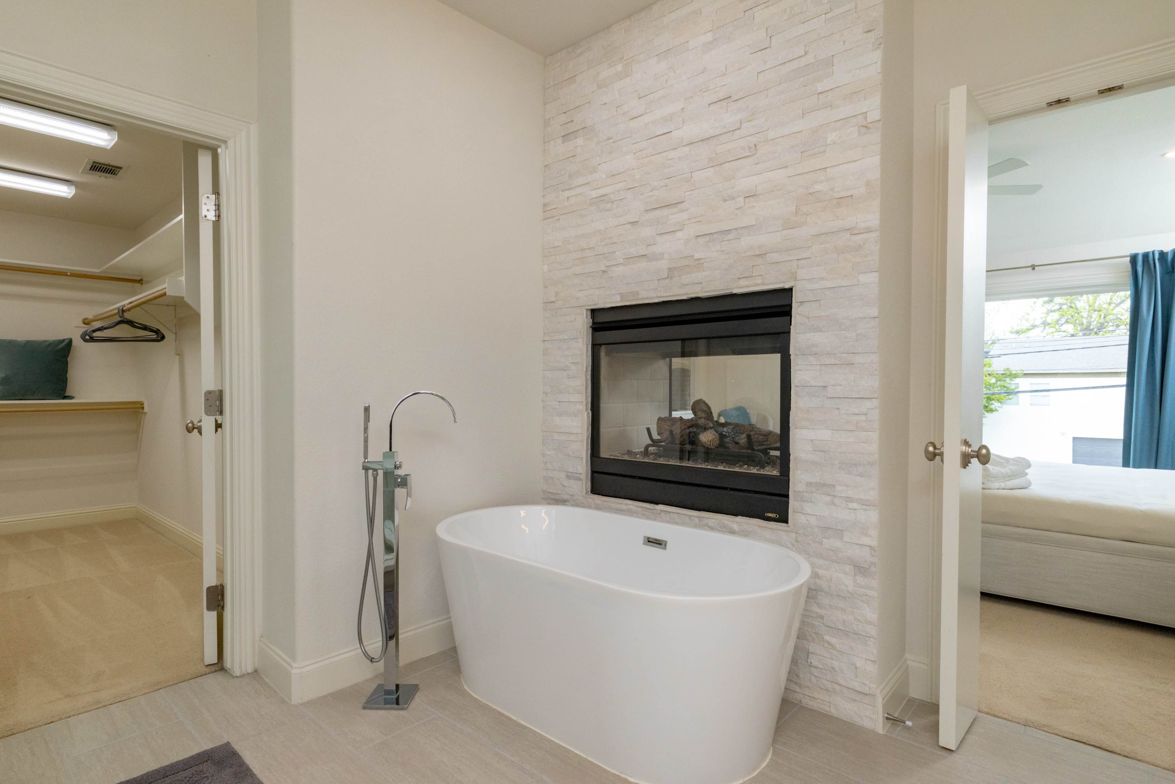 The image shows a modern bathroom featuring a freestanding bathtub next to a stone fireplace, with a closet visible in the background.