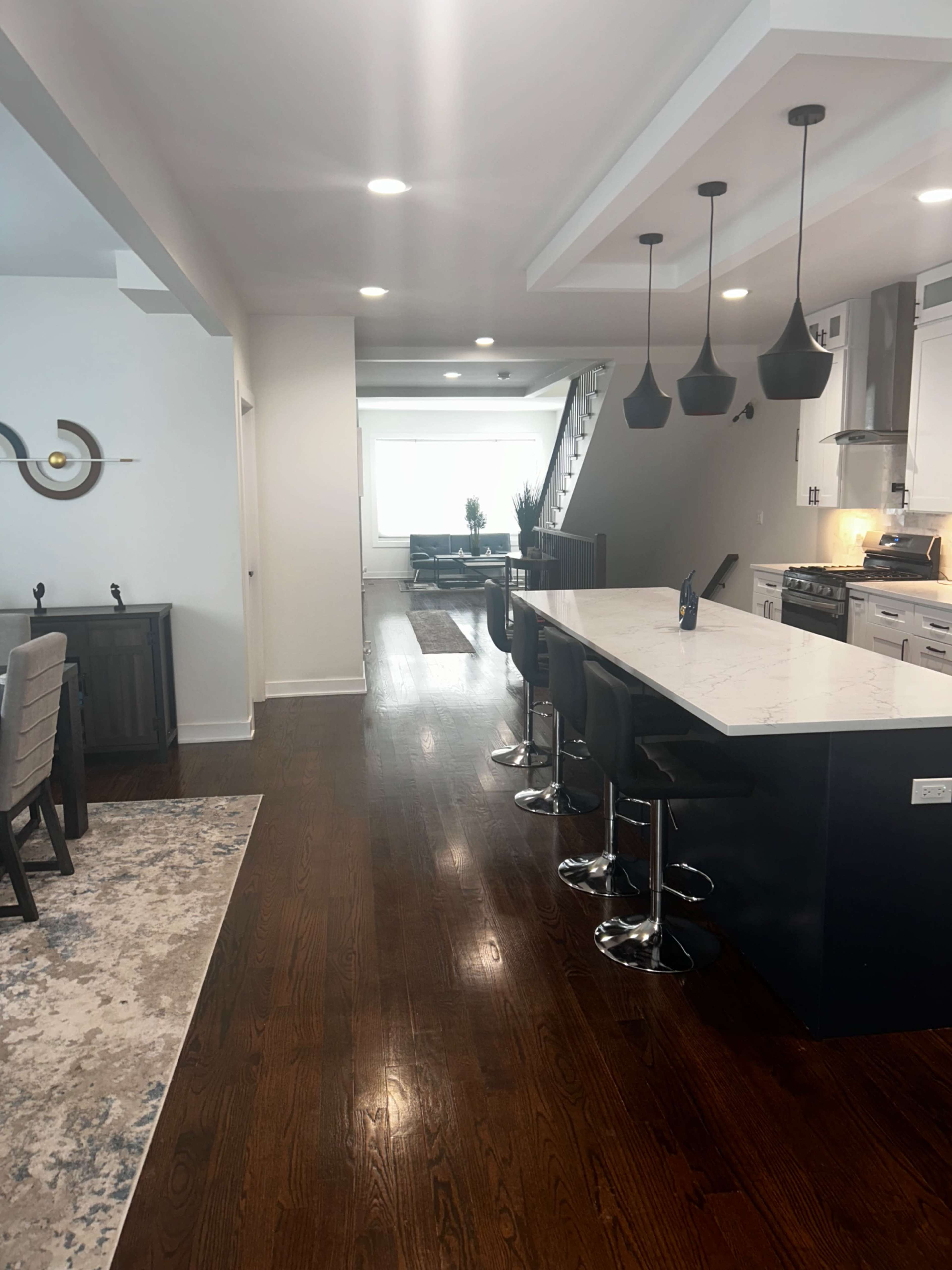 The image shows a modern kitchen and dining area with a large island, bar stools, and a staircase in the background.