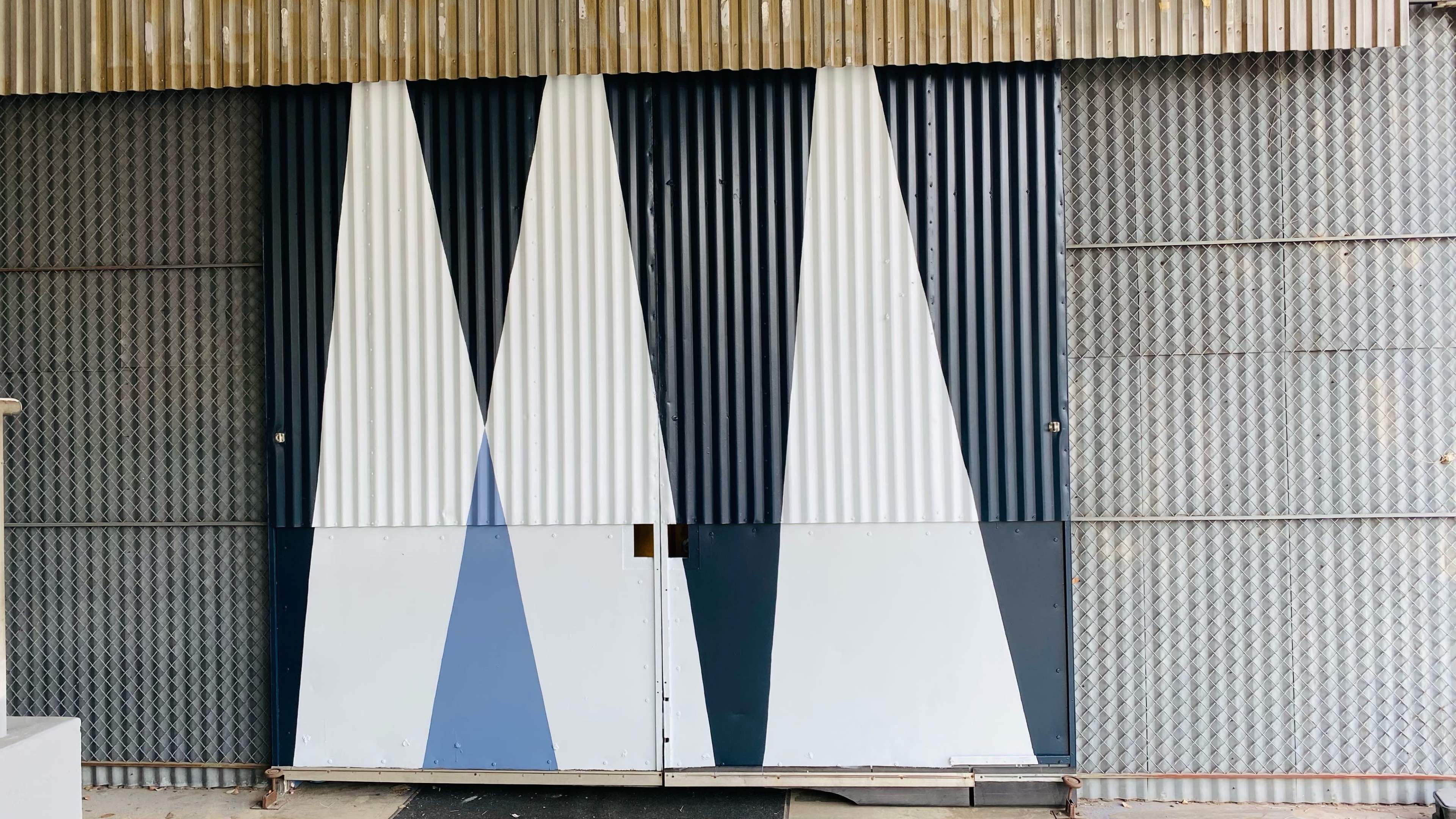 The image shows a multi-colored corrugated metal door with triangular patterns in shades of white, gray, and blue, set against a backdrop of wooden slats and textured metal.