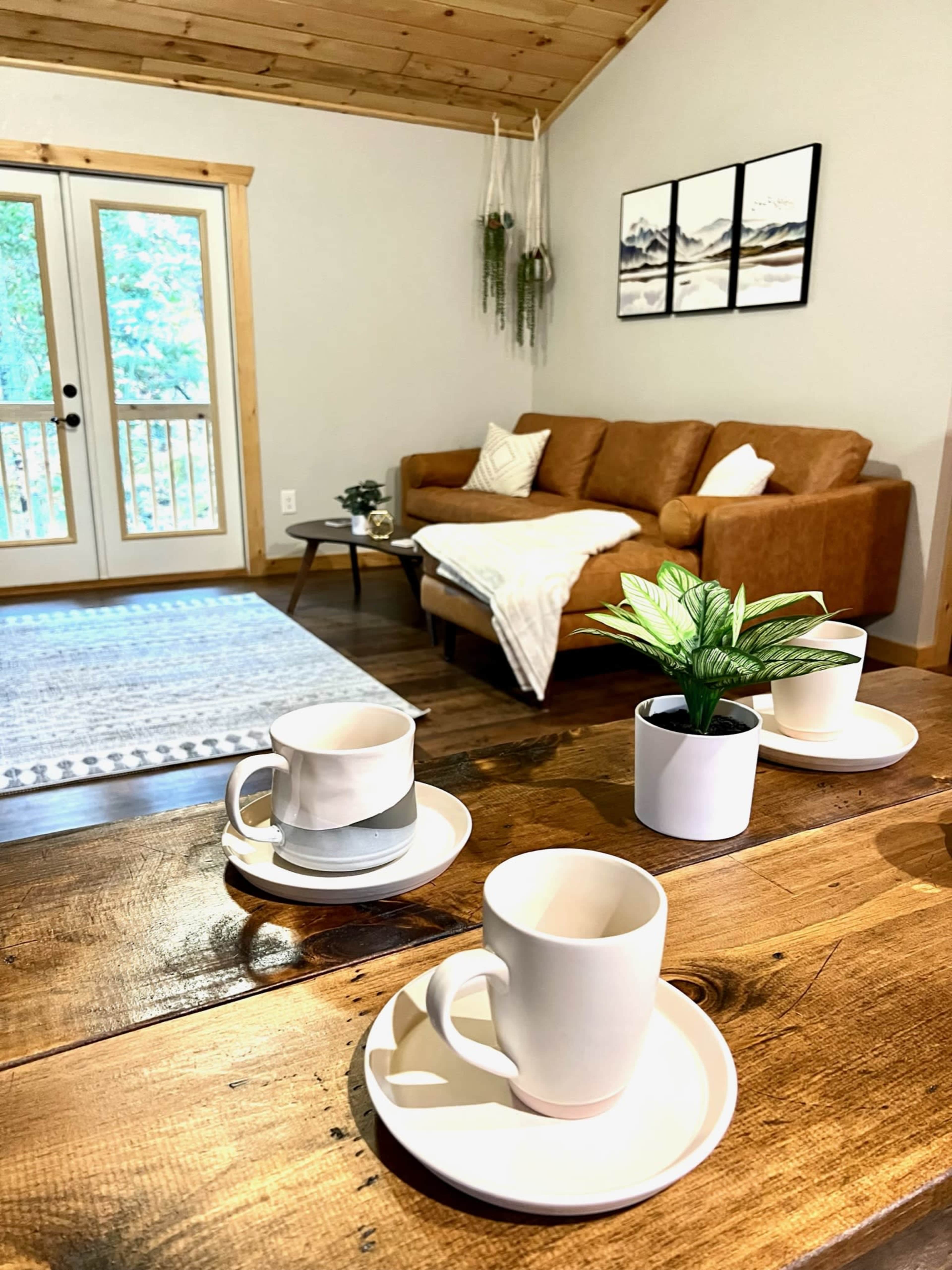 A coffee table with three cups and saucers is set in a cozy living room featuring a brown sofa, potted plant, and double doors leading to a deck.