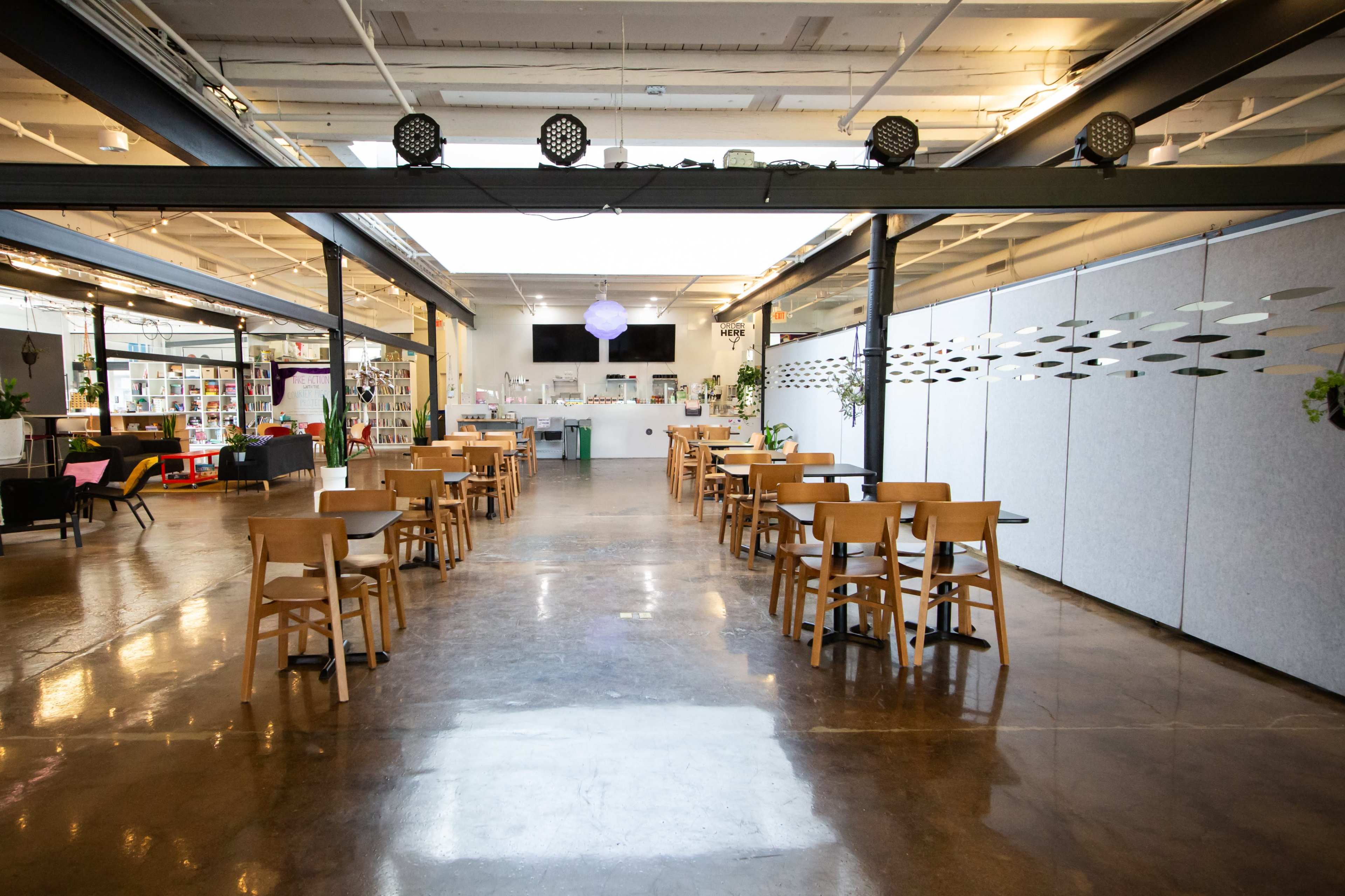 The image shows a spacious, well-lit interior featuring several wooden tables and chairs arranged in rows, with dividers and a counter area in the background.