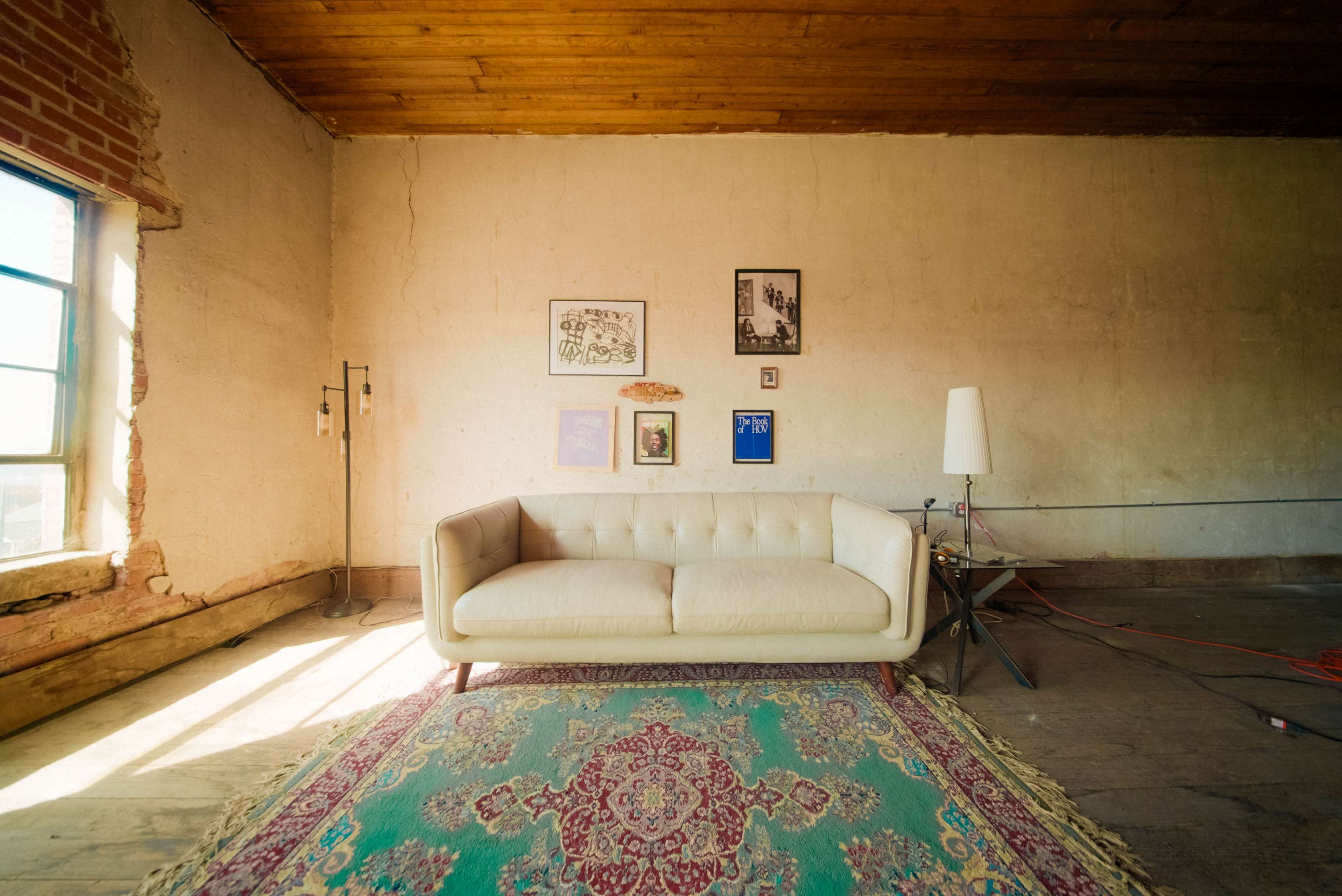 The image shows a minimalist room with a light-colored sofa, a patterned rug, and framed artwork on the exposed brick wall.