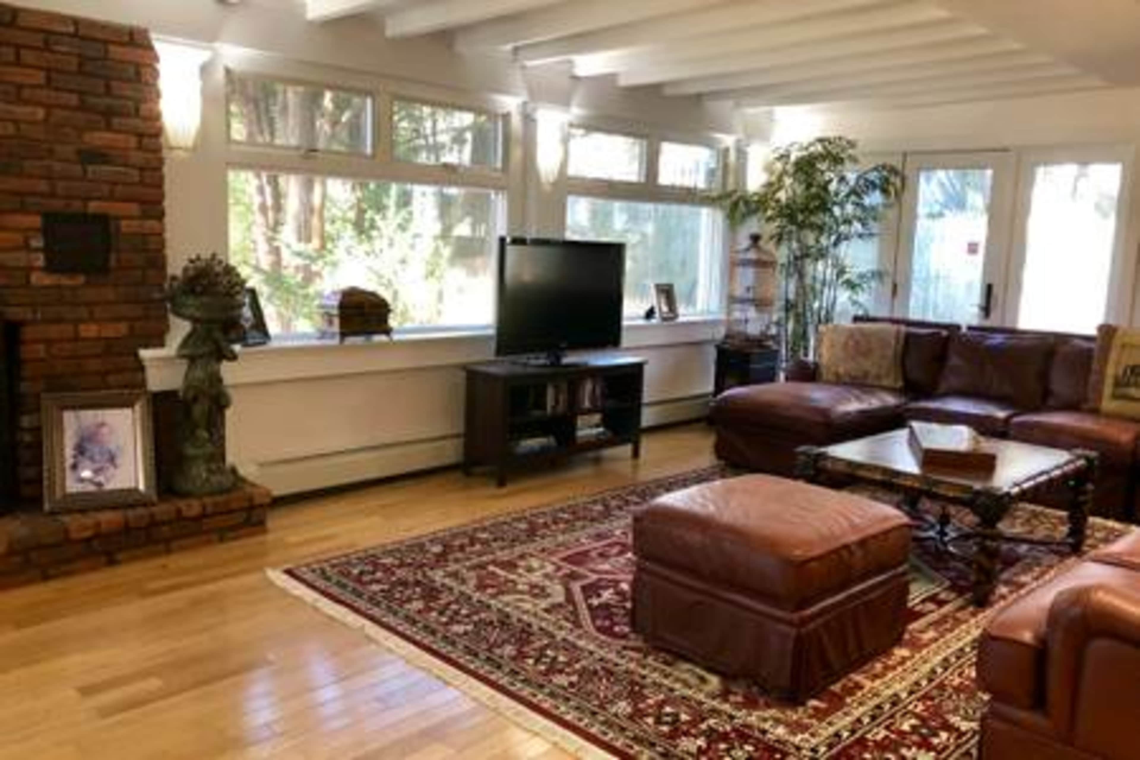 The image depicts a cozy living room with large windows, a television, a brown leather sectional sofa, and a decorative rug atop wooden flooring.