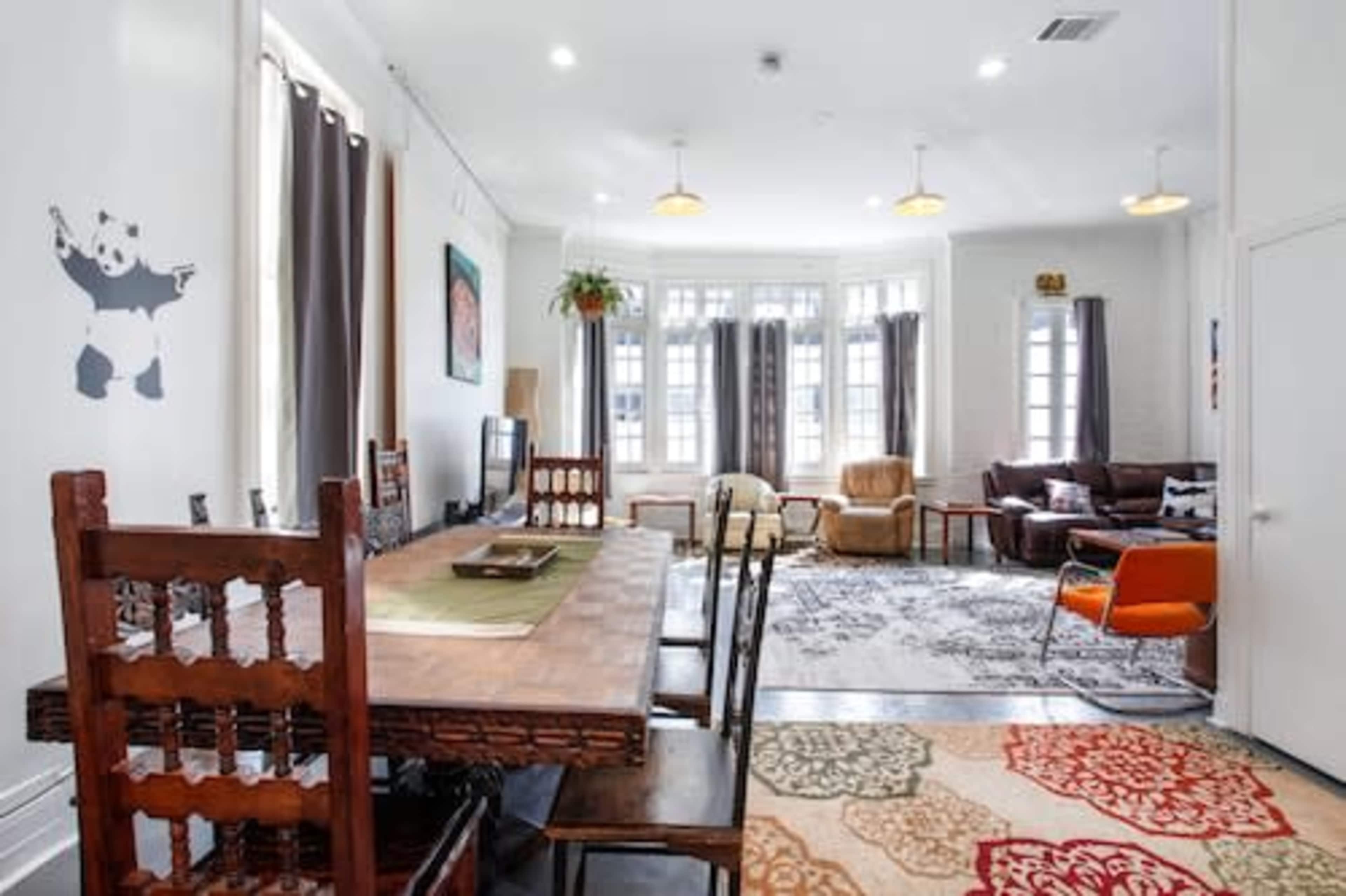 A spacious, well-lit living area featuring a dining table with chairs, a television, a cozy seating arrangement, and large windows.
