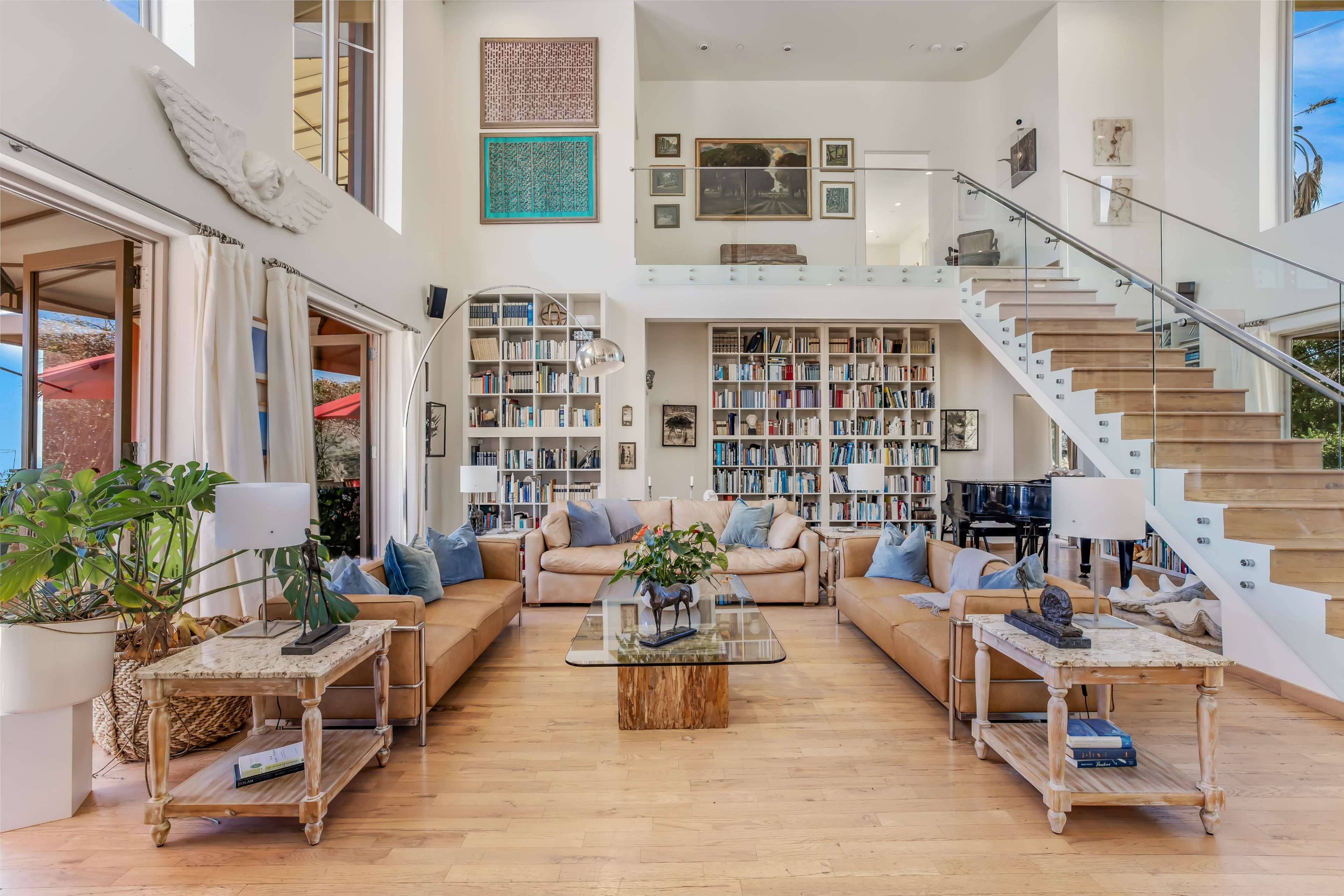 A spacious living room with two large sofas arranged around a glass coffee table, a bookshelf filled with books, and a staircase leading to an upper level.
