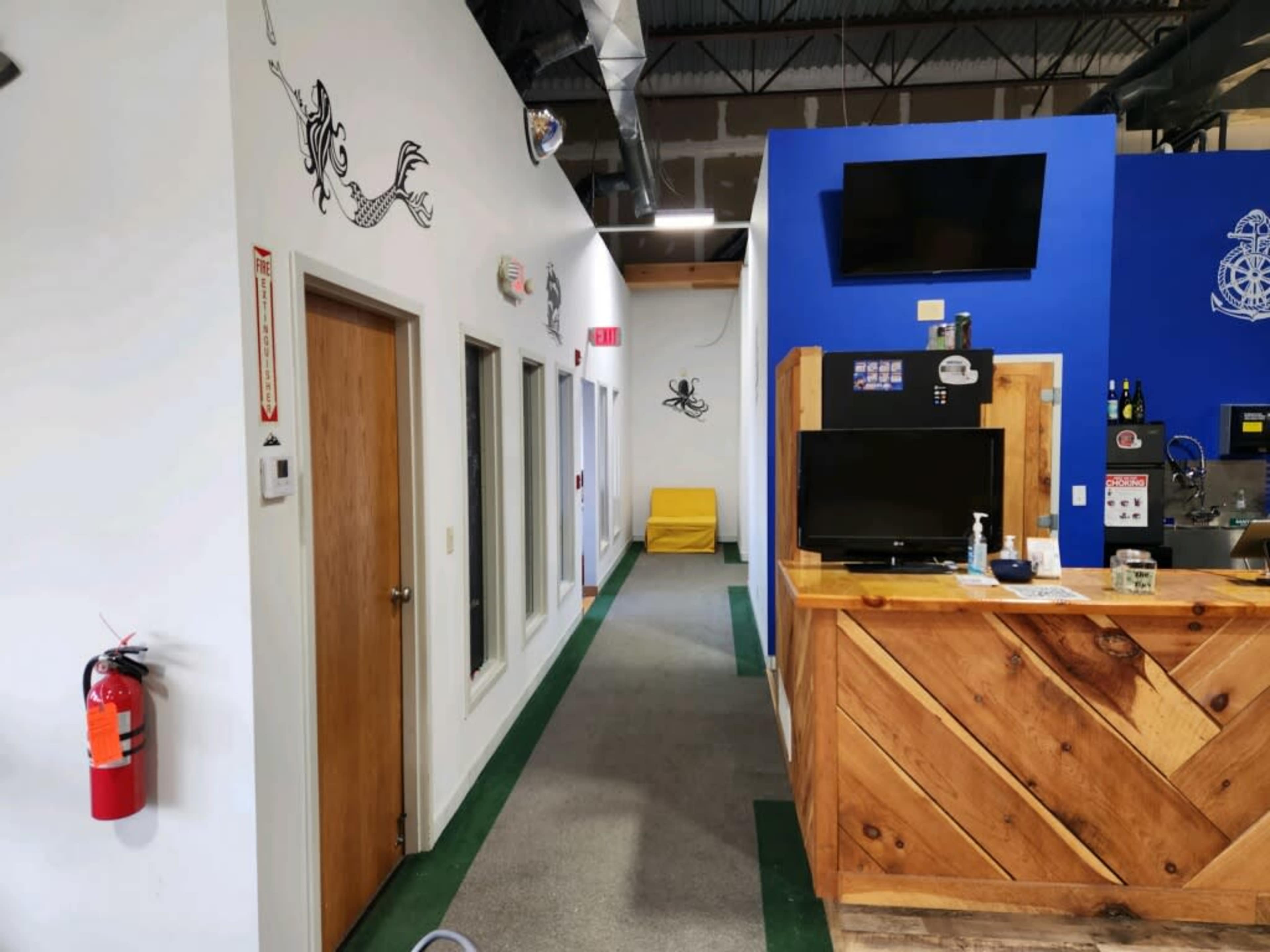 A well-lit indoor space with a wooden reception area on the right, a hallway with doors on the left, and a blue wall featuring a television and decor.