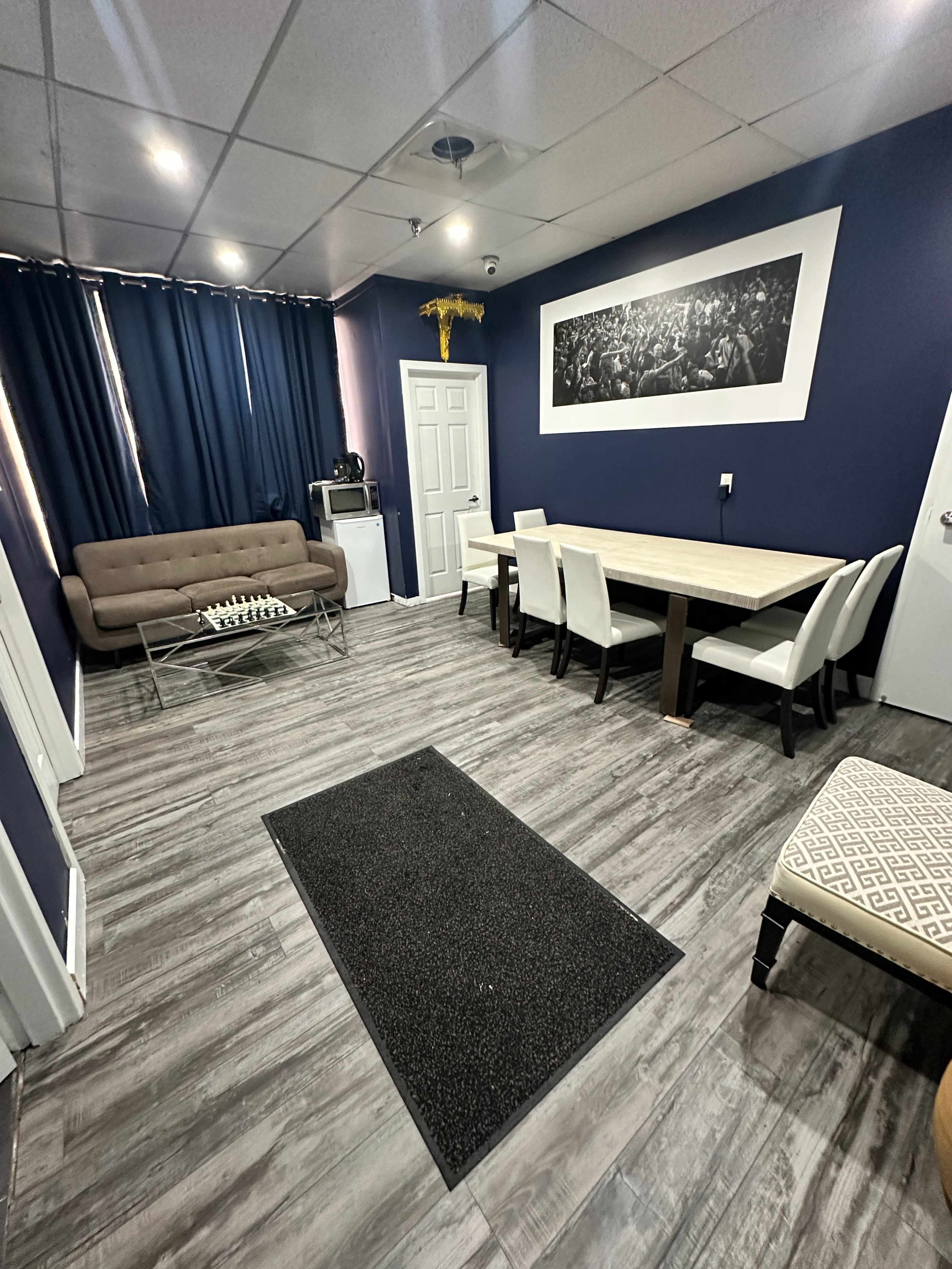 The image shows a room featuring a seating area with a sofa, a dining table with chairs, and dark blue walls.