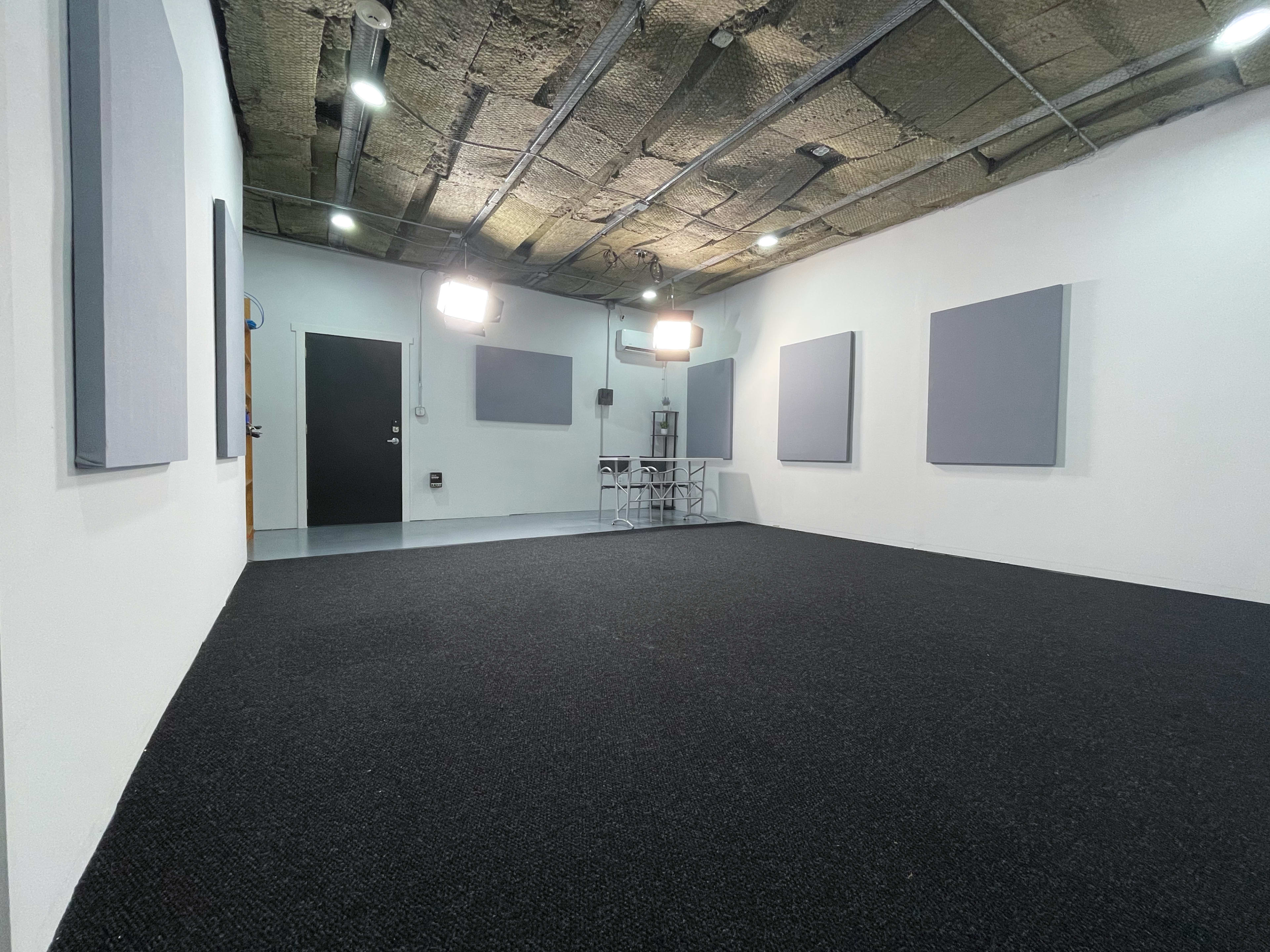 The image shows a soundproofed studio space with grey acoustic panels on the walls, a black carpeted floor, and a table with chairs in the center.