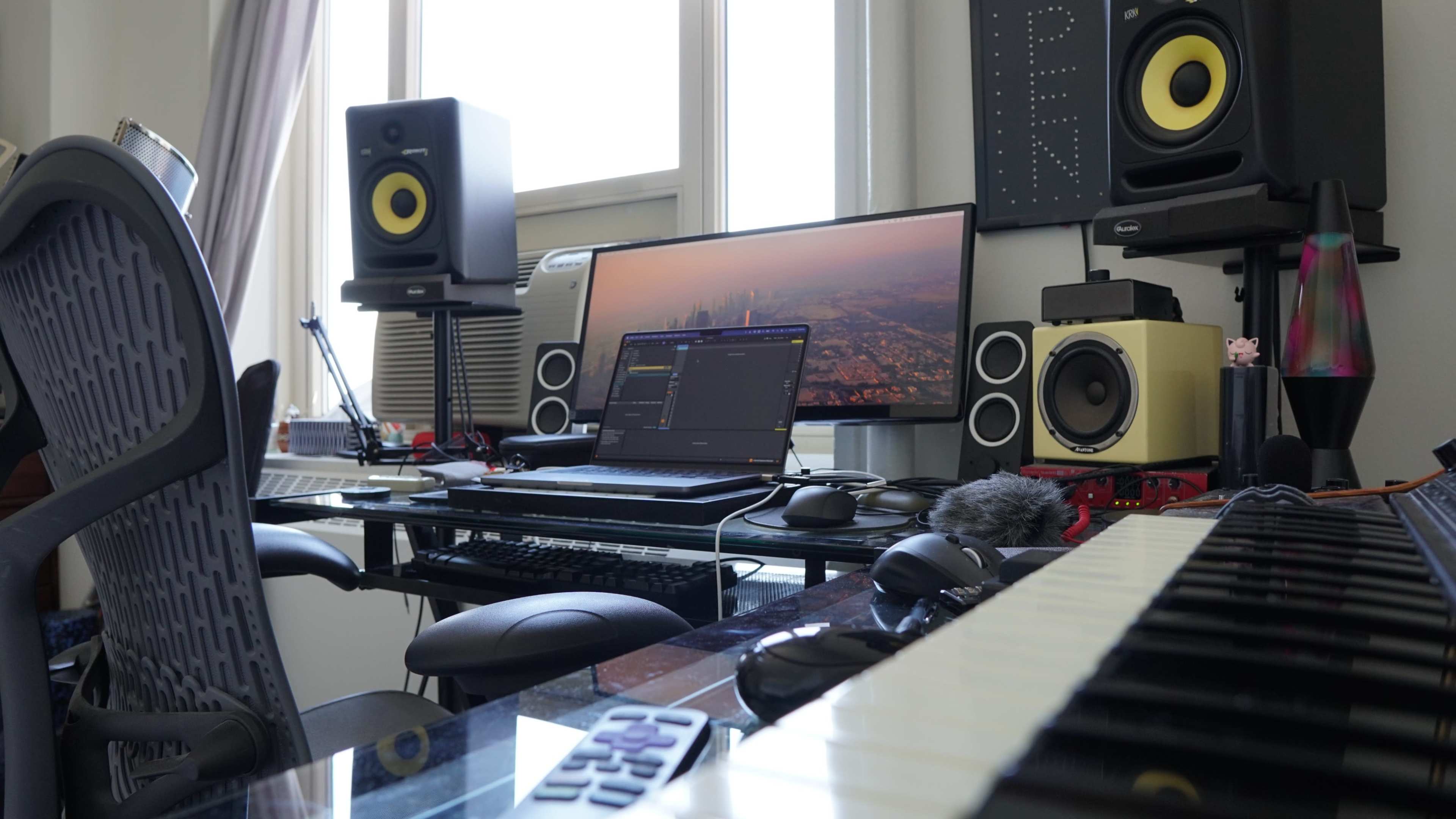The image shows a music production workspace featuring a computer with dual monitors, studio speakers, a keyboard, and various audio equipment on a glass desk.