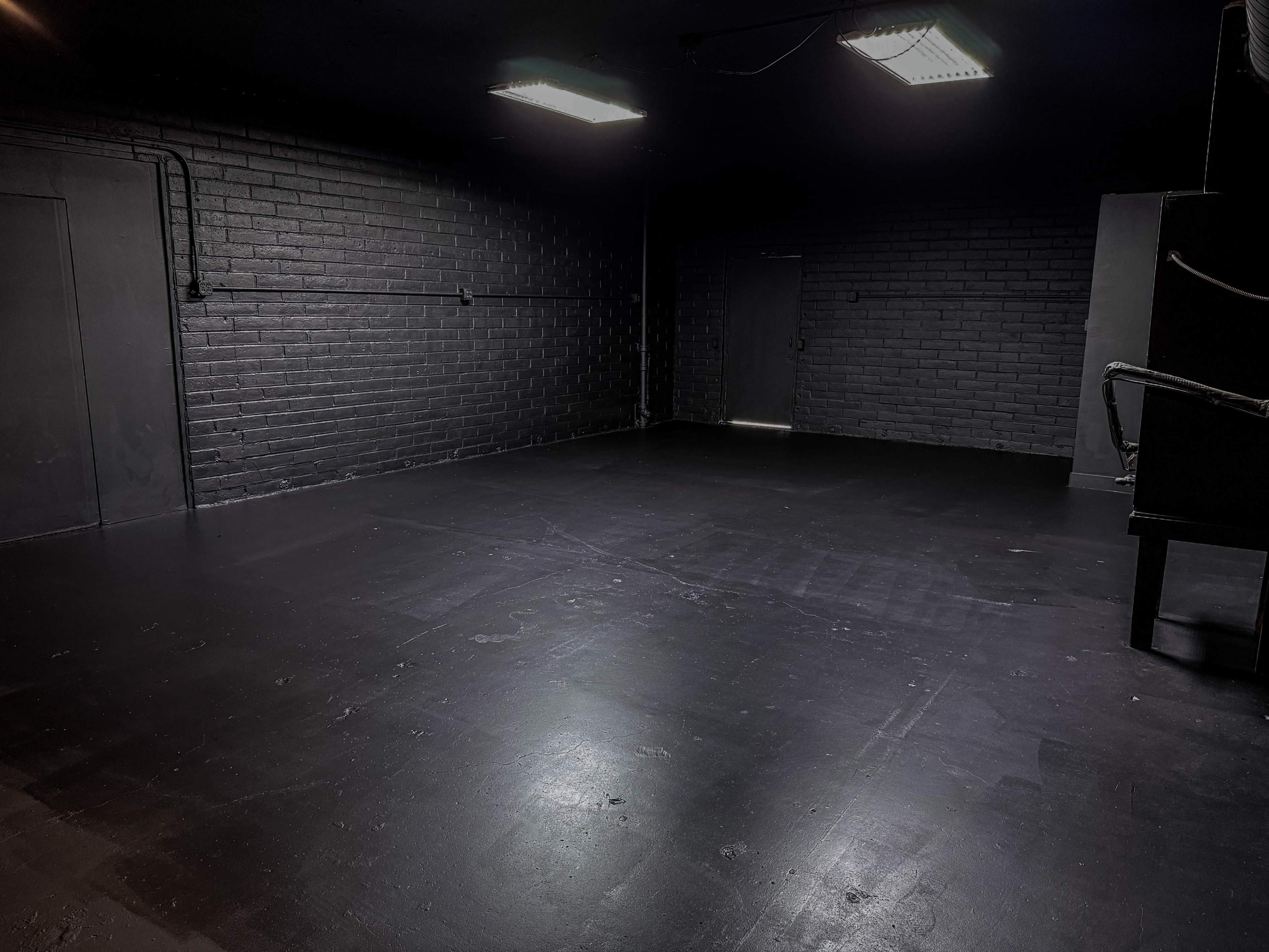 The image shows a sparsely furnished black room with brick walls, a smooth dark floor, and two overhead lights illuminating a doorway at the far end.