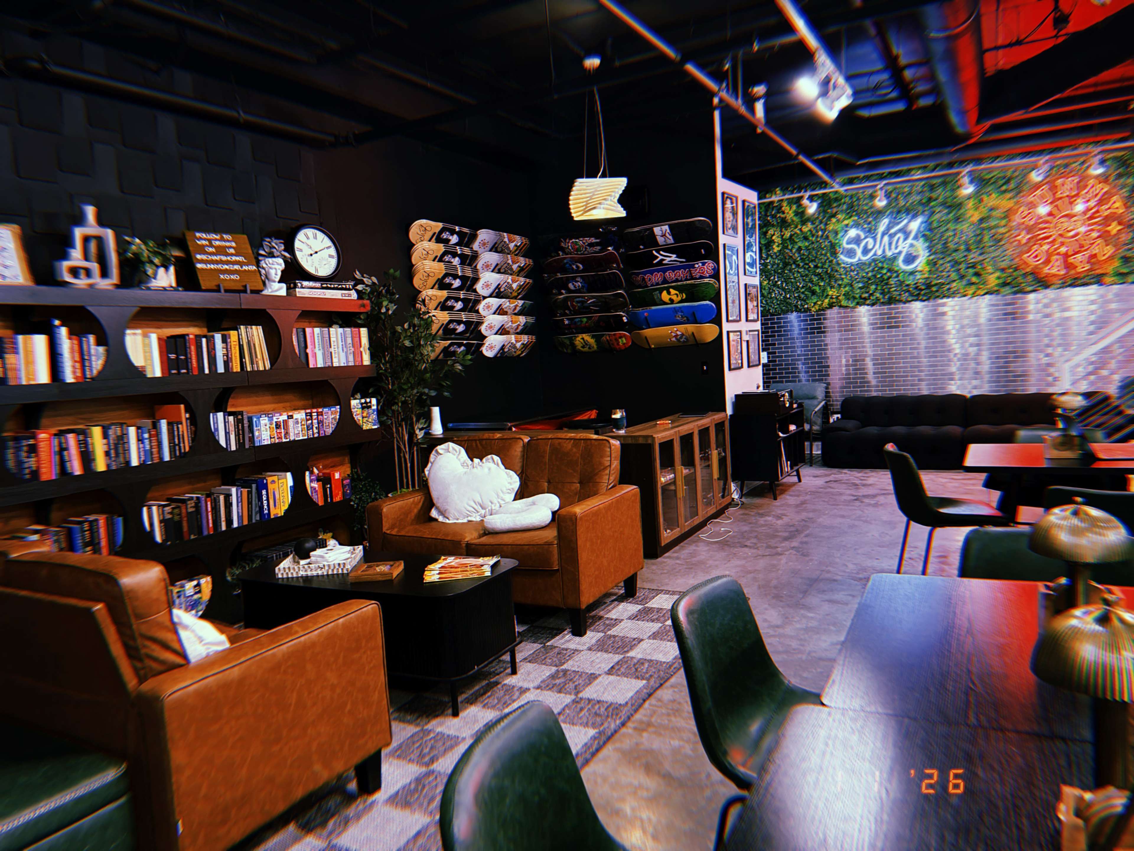 The image shows a cozy café interior with brown armchairs, wooden bookshelves filled with books, skateboards mounted on the wall, and a green wall adorned with plants.