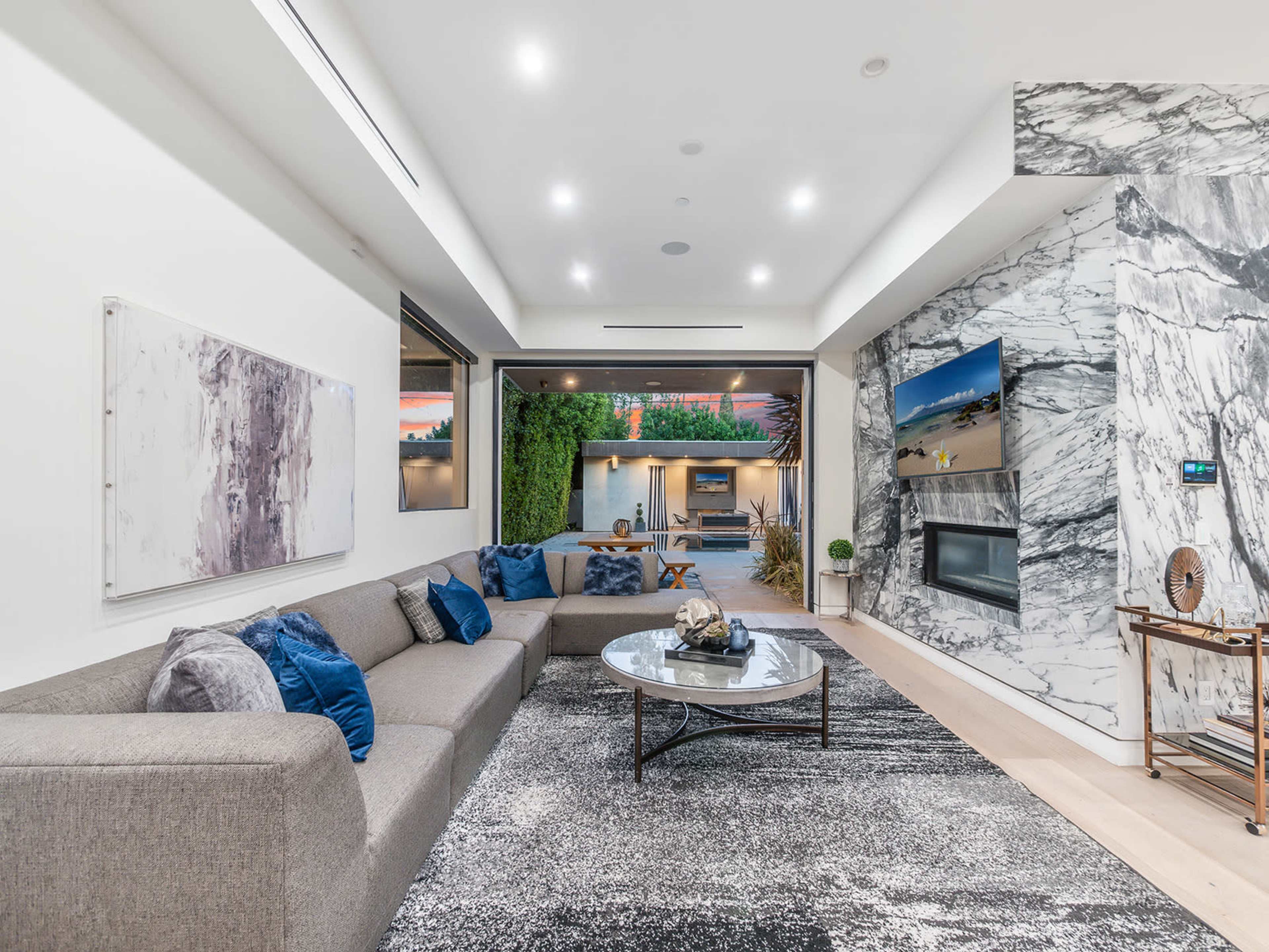 A modern living room featuring a large gray sectional sofa, a round coffee table, and a decorative wall with marble accents, with large windows providing a view of the outdoors.