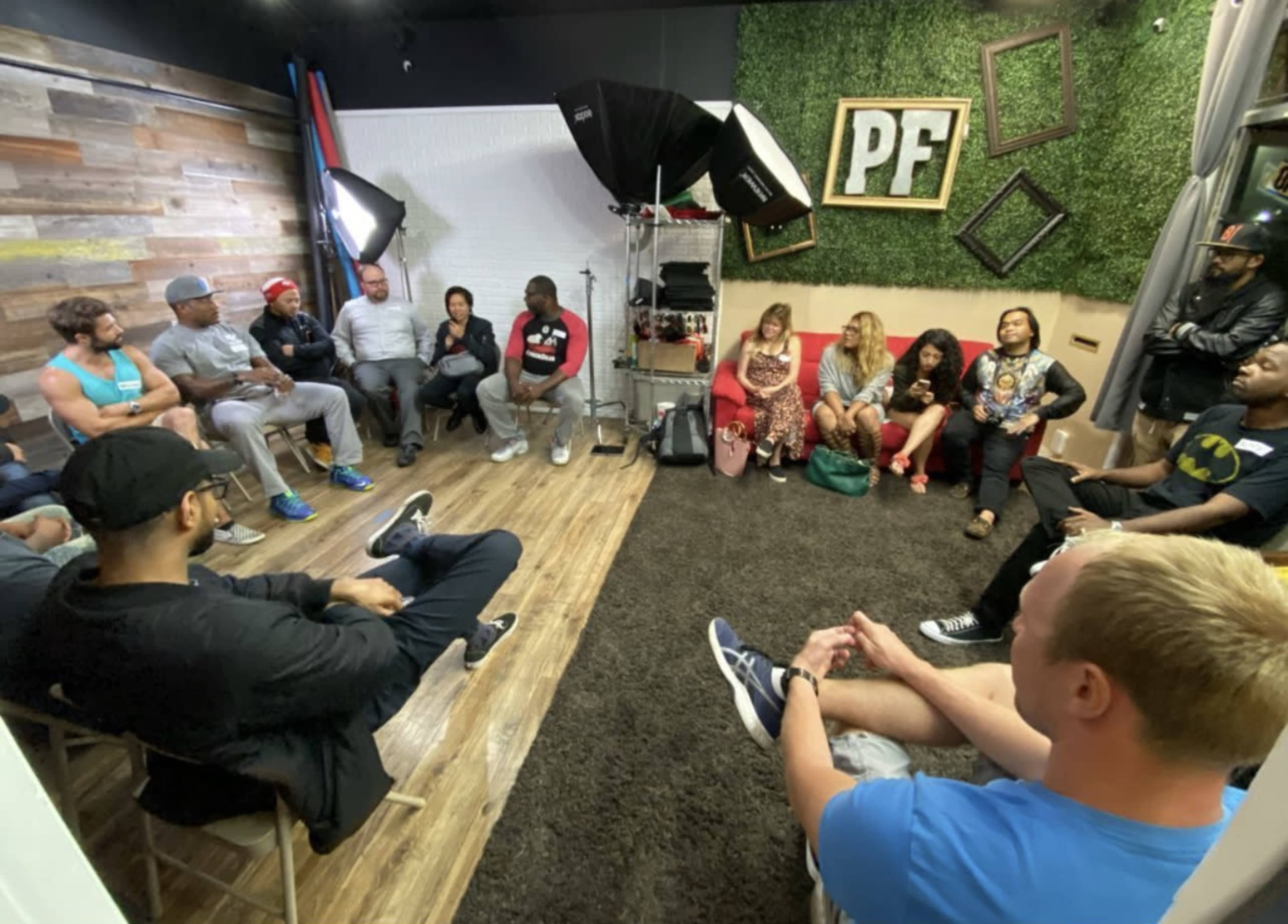 A diverse group of people is seated in a circle in a well-lit room with a green wall, discussing in a casual setting.