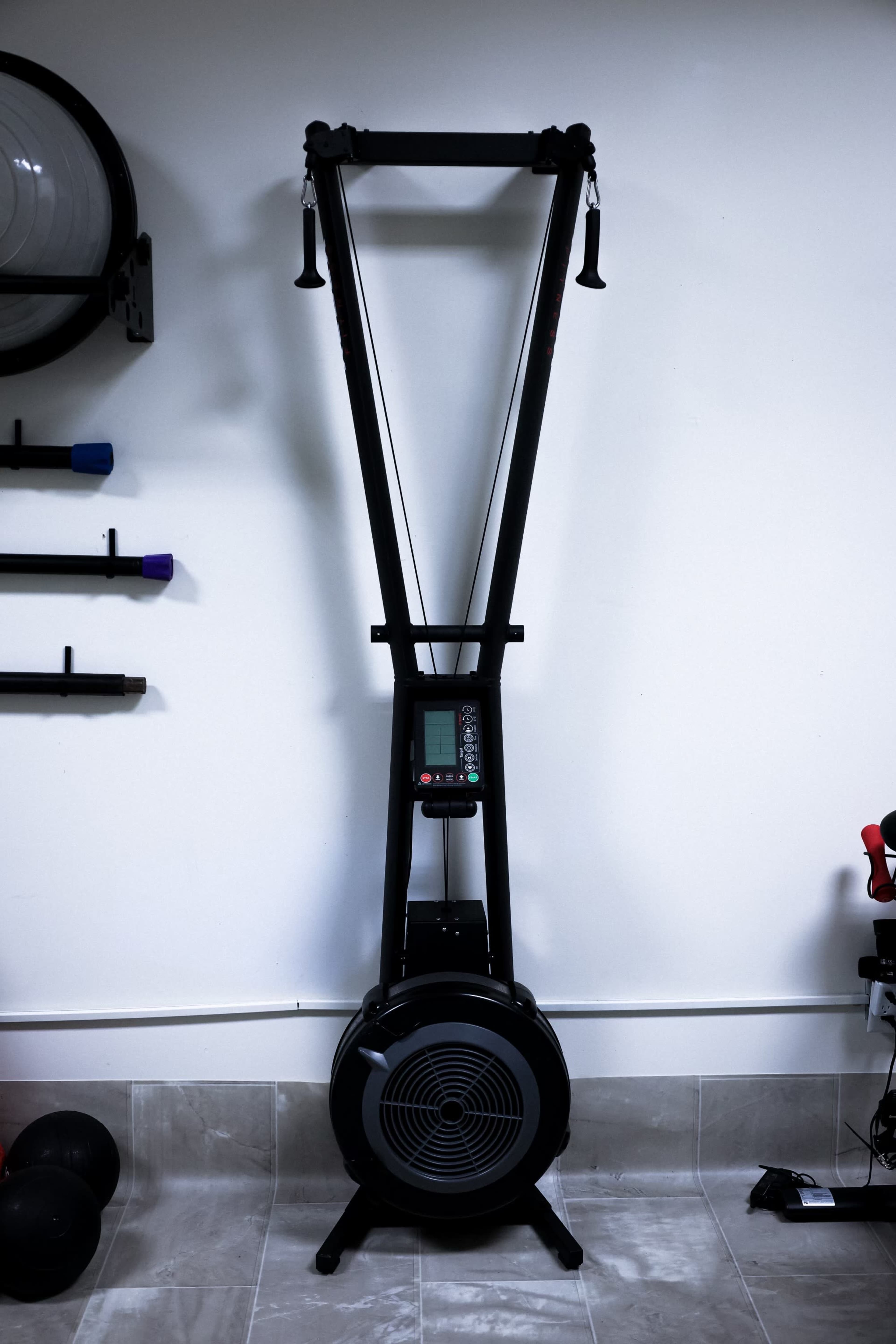 A vertical rowing machine with a digital display, positioned against a plain wall in a fitness space.