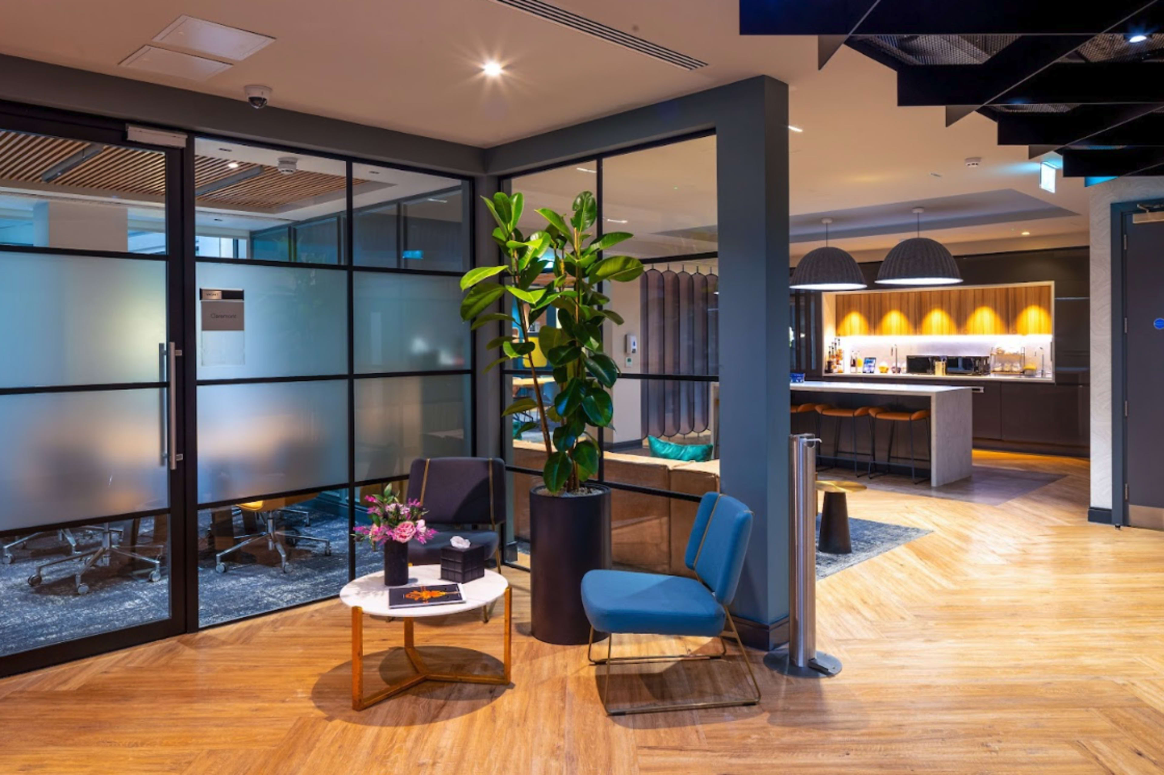 A modern office lobby featuring a small table with flowers, two chairs, and a glass partition leading to a conference room.