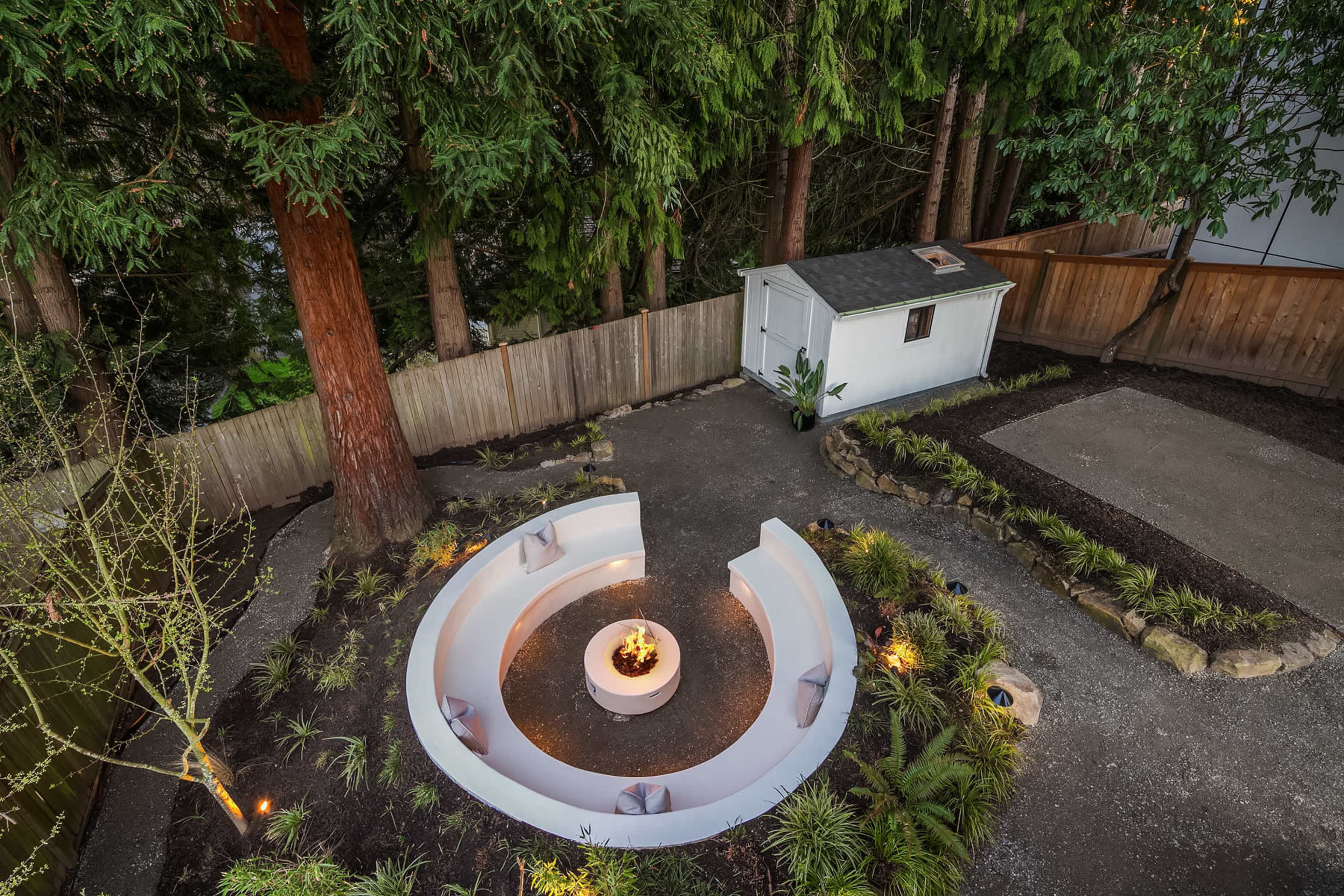 The image shows a circular fire pit area surrounded by seating, with a small shed and landscaped garden elements in a backyard setting.