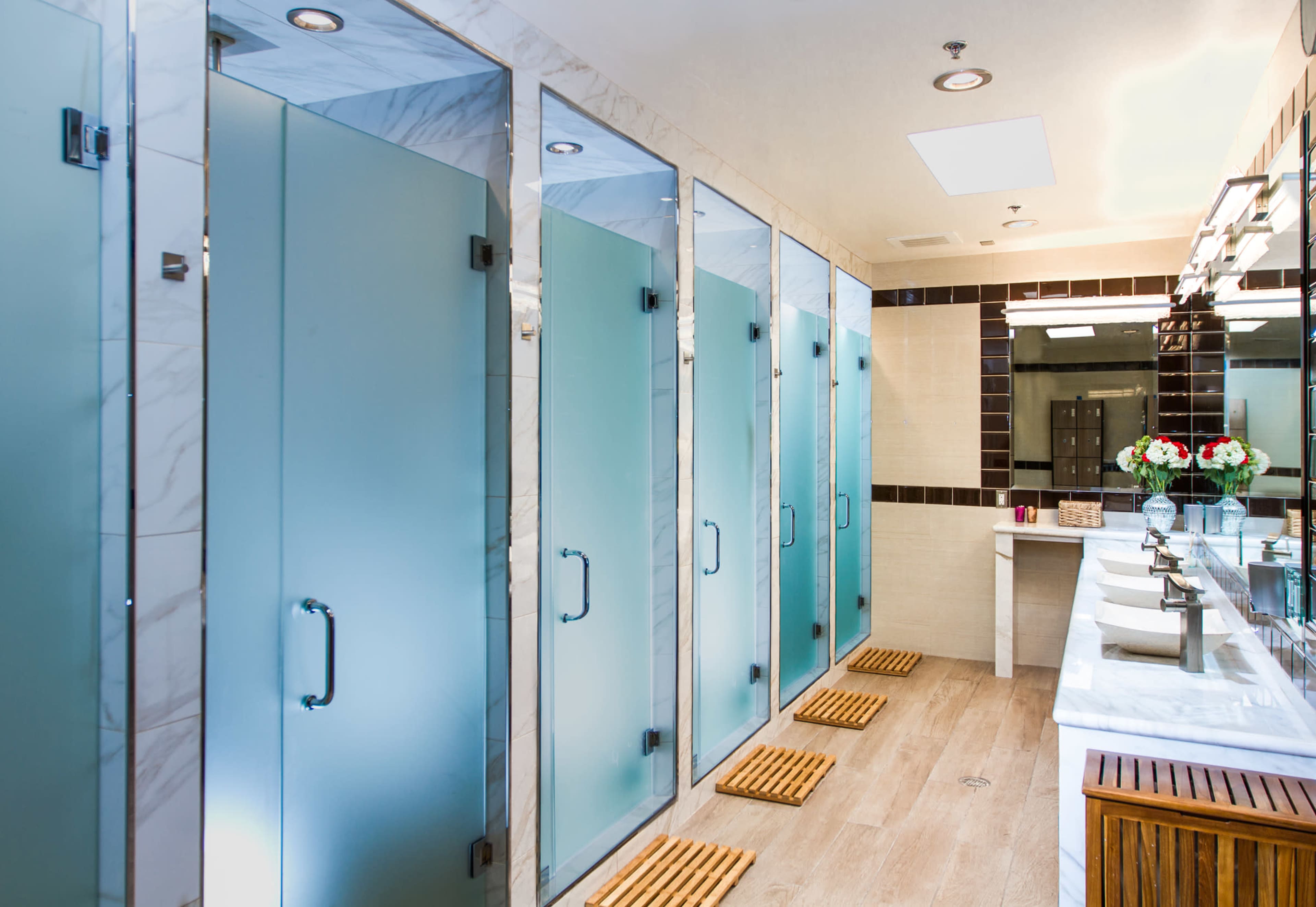 The image shows a modern bathroom with several frosted glass shower stalls, wooden mats, and a double sink vanity with decorative flowers.