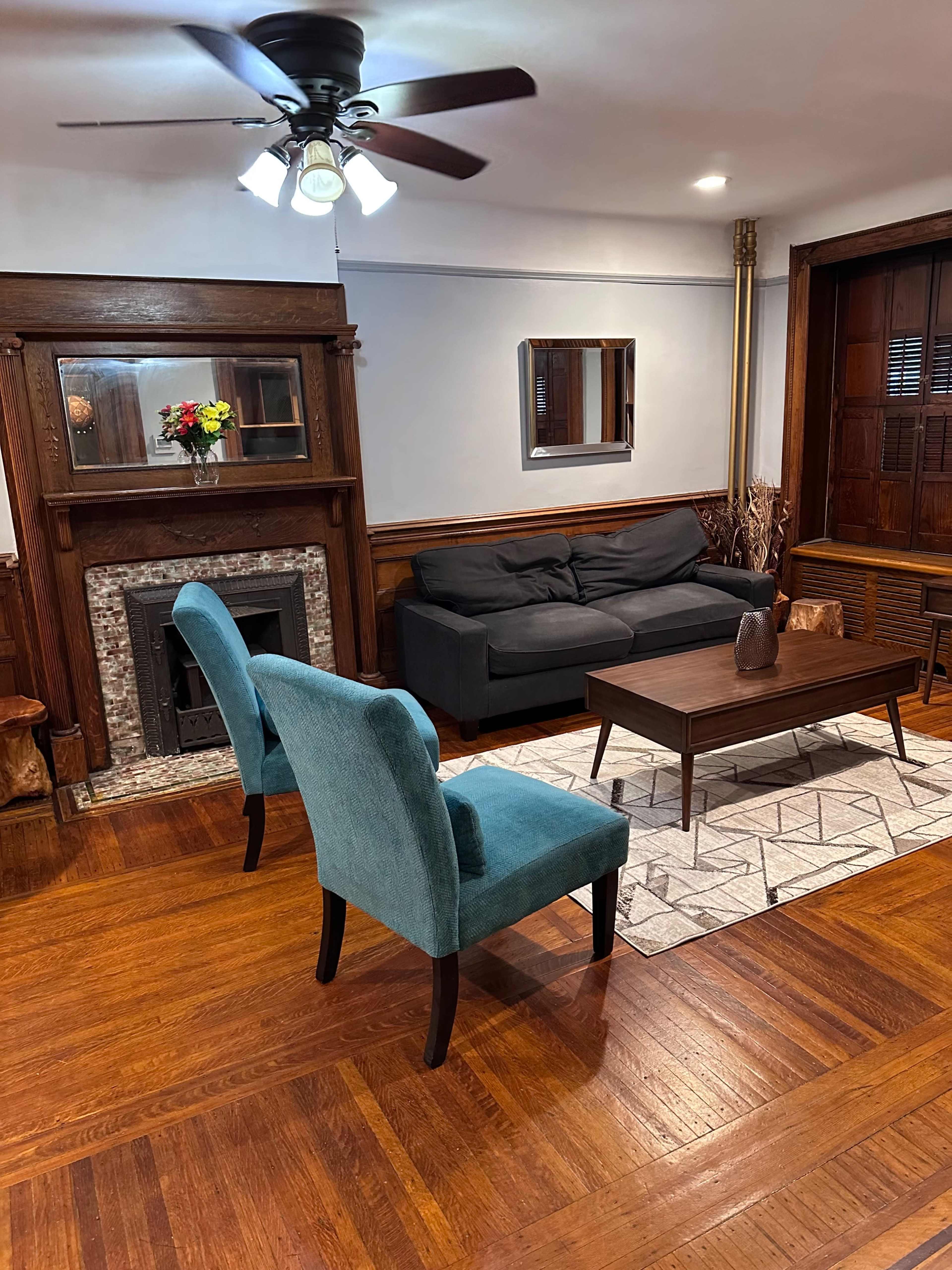 The image shows a living room with a black sofa, a wooden coffee table, two teal chairs, a decorative rug, and a fireplace with ornate woodwork.