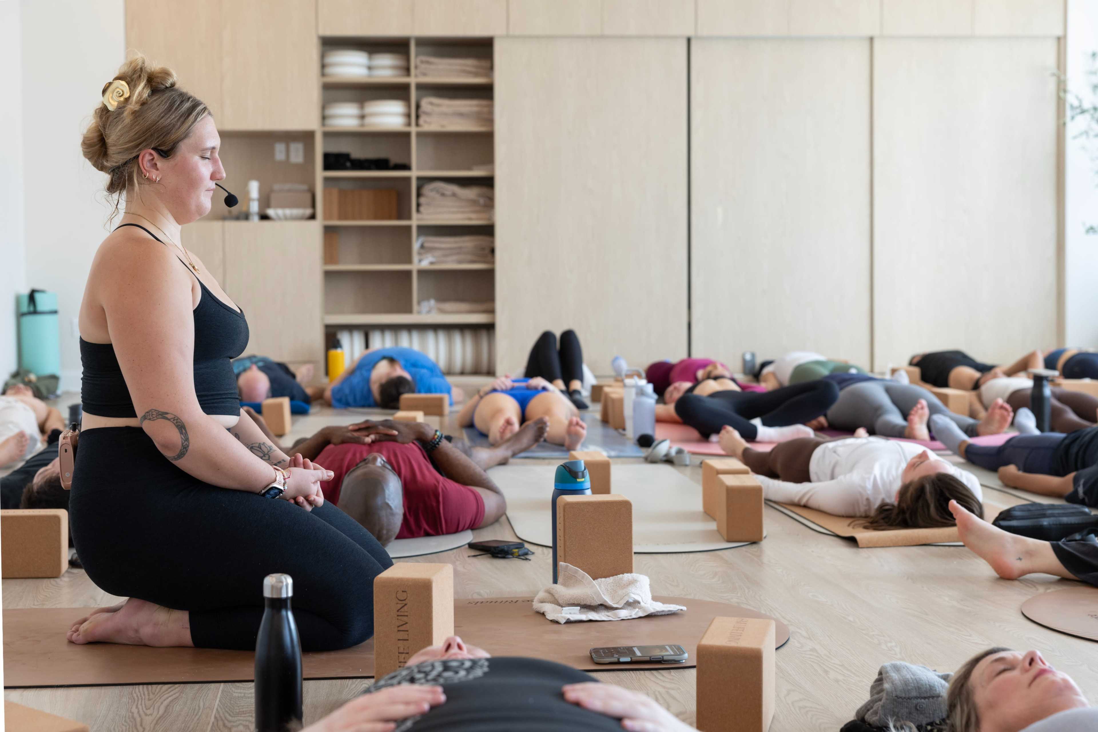 A yoga instructor sits on a mat while participants lie on the floor in a studio during a class.