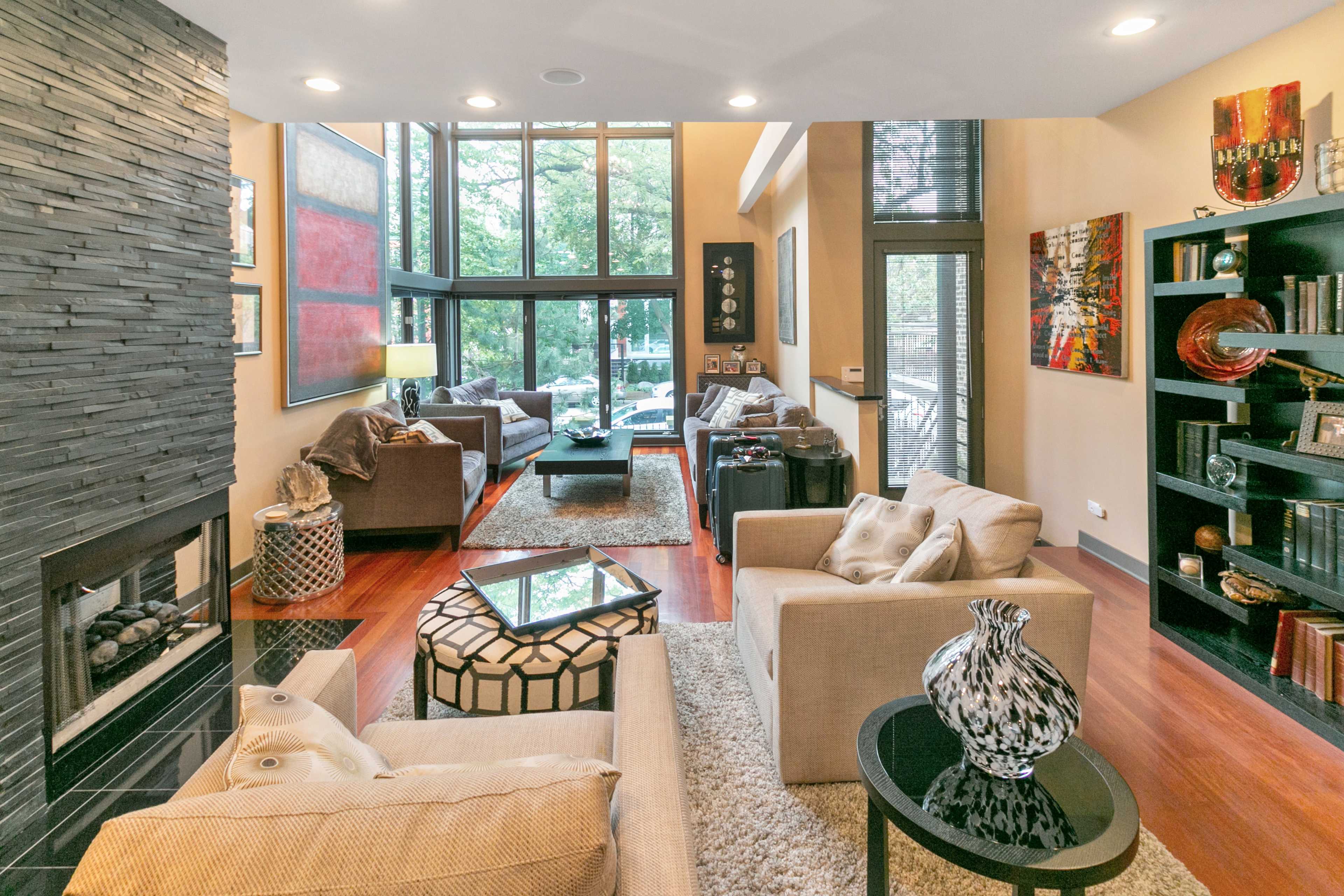 A modern living room features large windows, a stone fireplace, and comfortable seating arrangements with decorative elements.