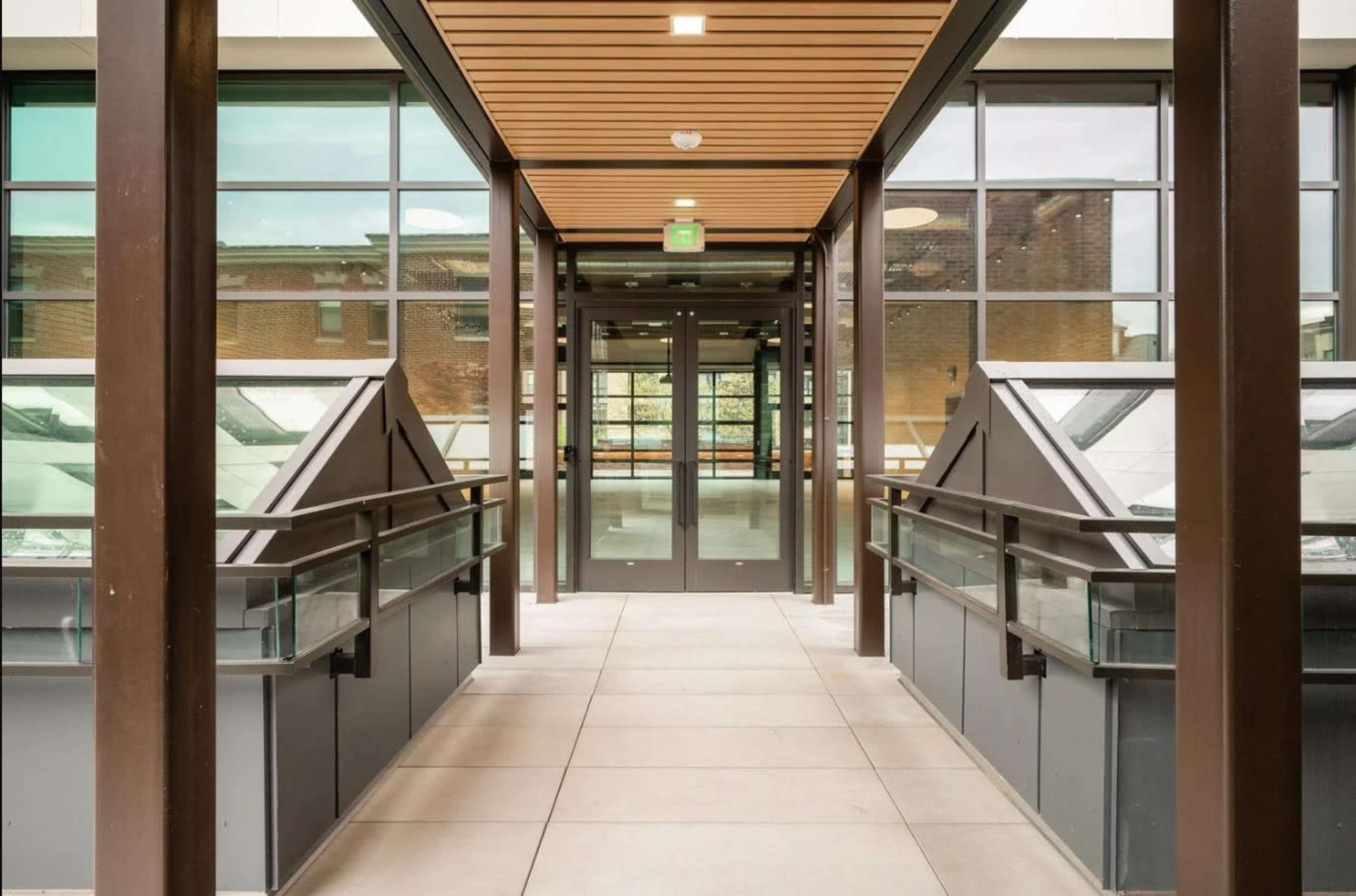 The image shows a contemporary hallway with large glass panels and a door at the end, framed by wooden ceiling slats.