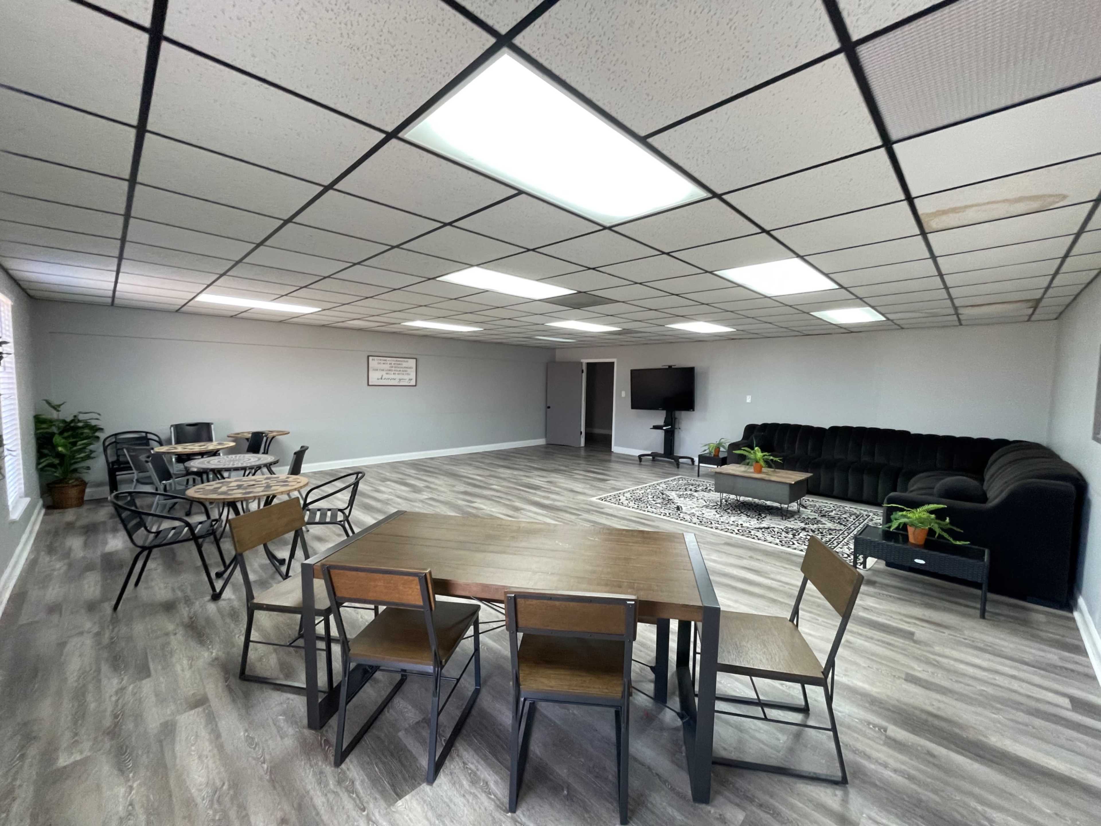 The image shows a spacious room with multiple tables and chairs, a black couch, and a TV set against the wall.