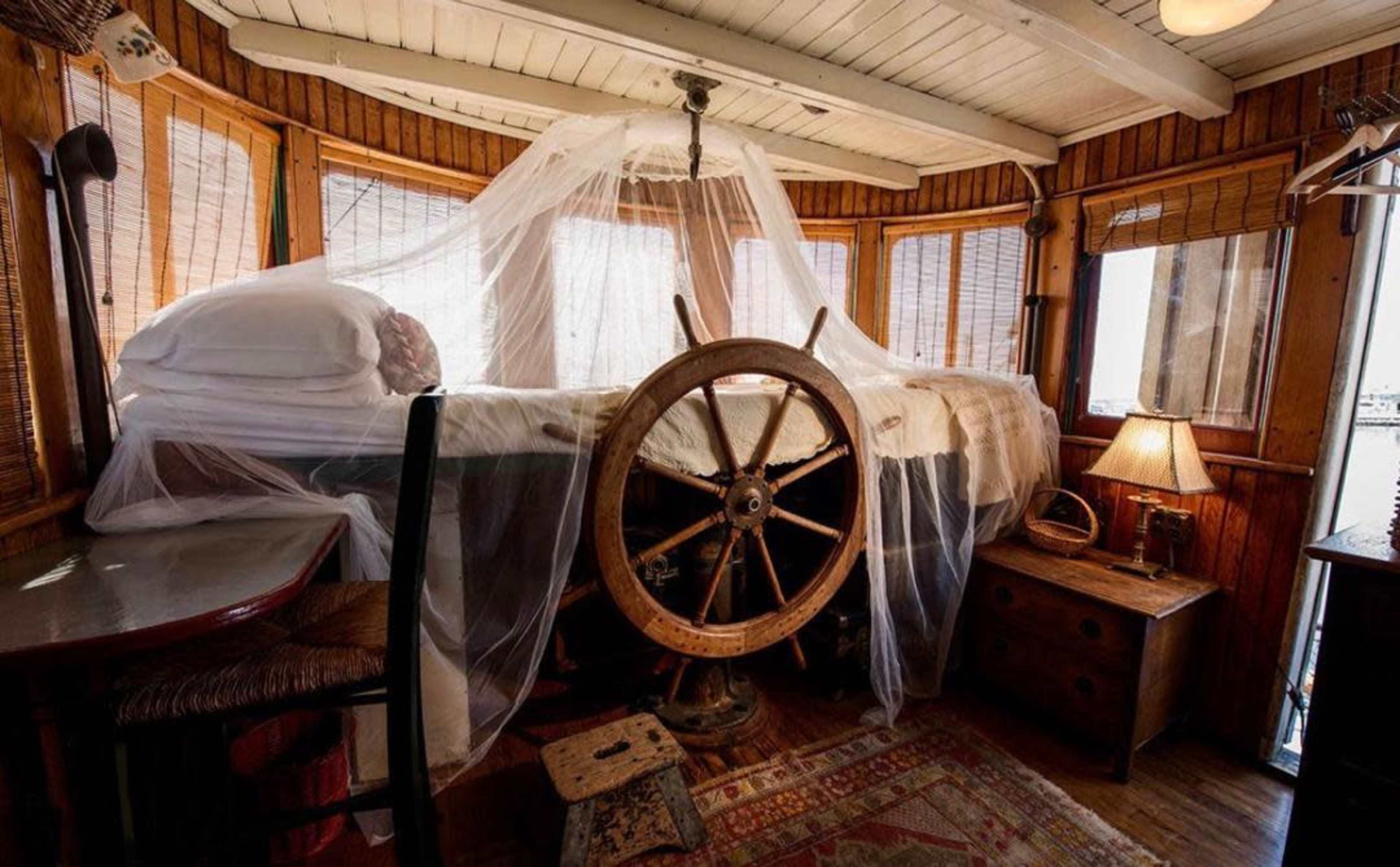 The scene shows a wooden room featuring a bed draped with a sheer canopy, a ship's wheel as a focal point, and rustic furniture including a lamp and a small table.