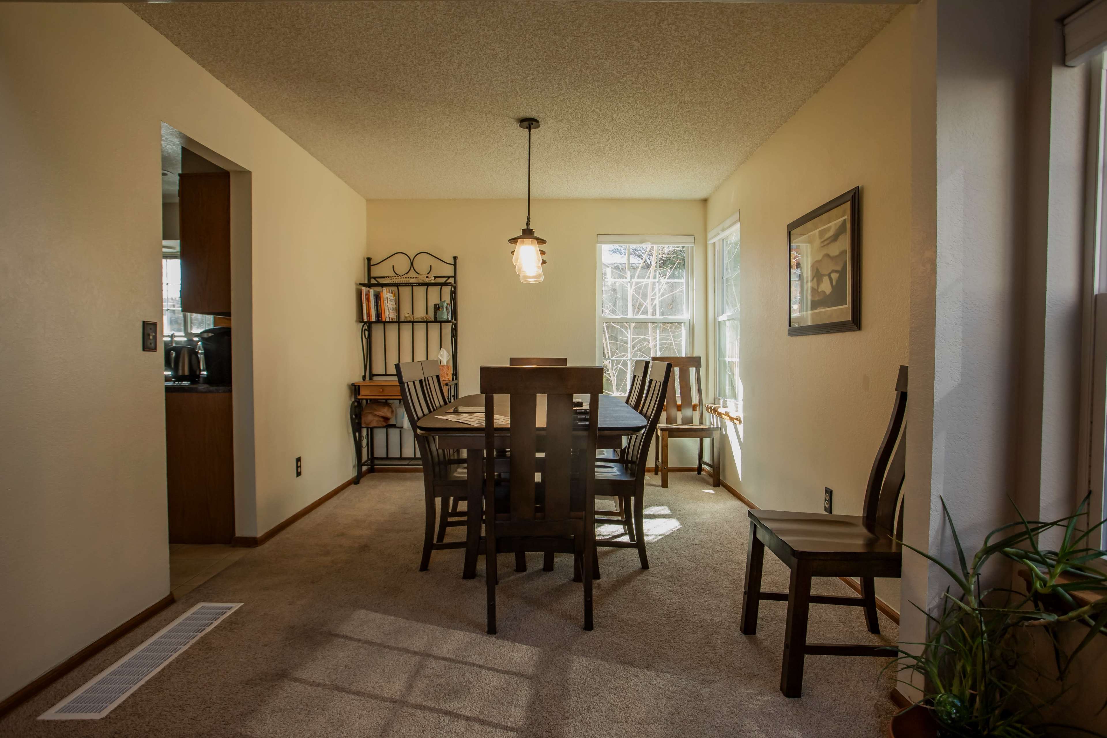 A dining room features a wooden table surrounded by chairs, with large windows allowing natural light and a small shelf against the wall.