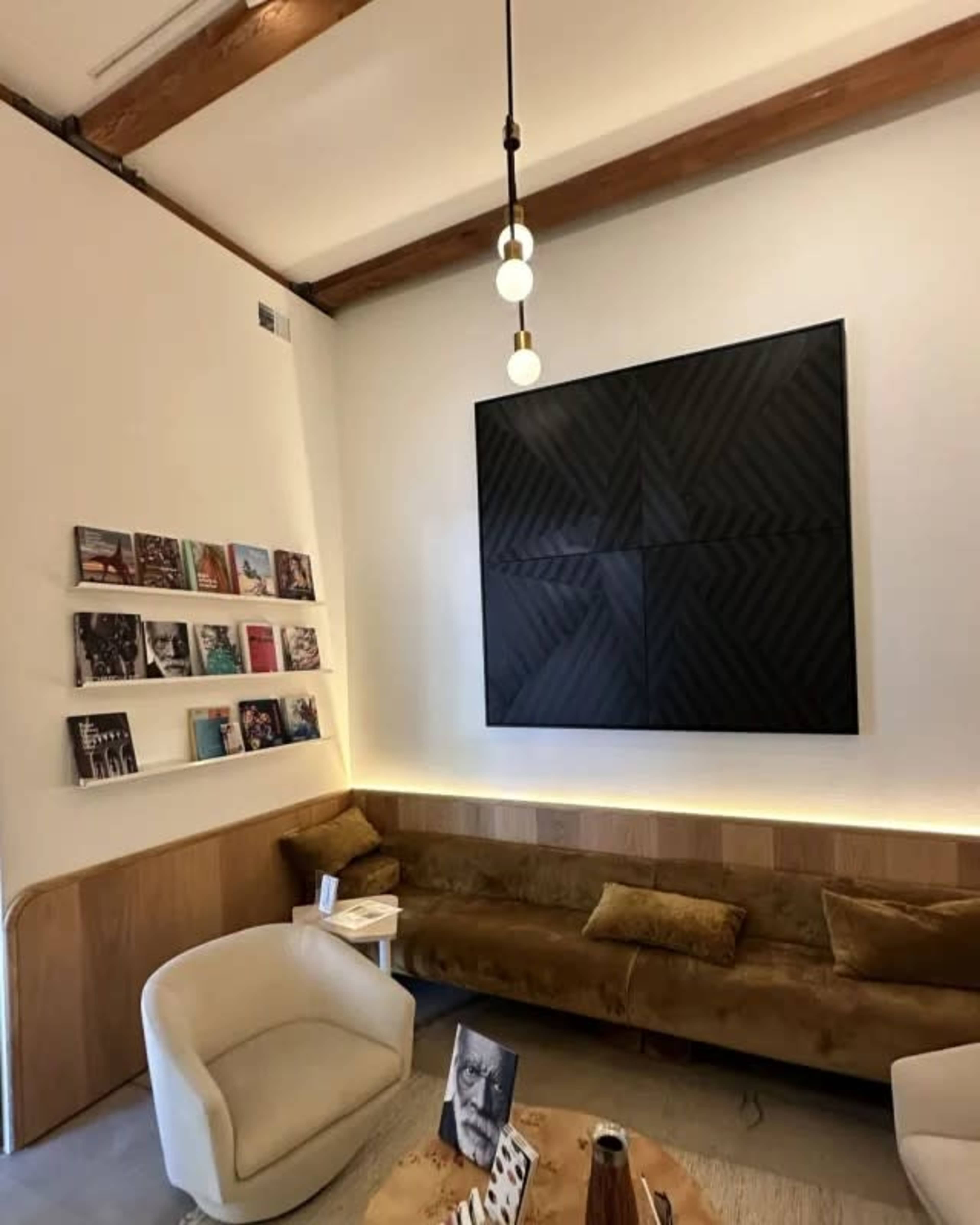 The image depicts a modern interior space featuring a low, brown couch, a circular white chair, a wooden table, and a large black artwork on the wall, along with a shelf displaying various magazines or books.