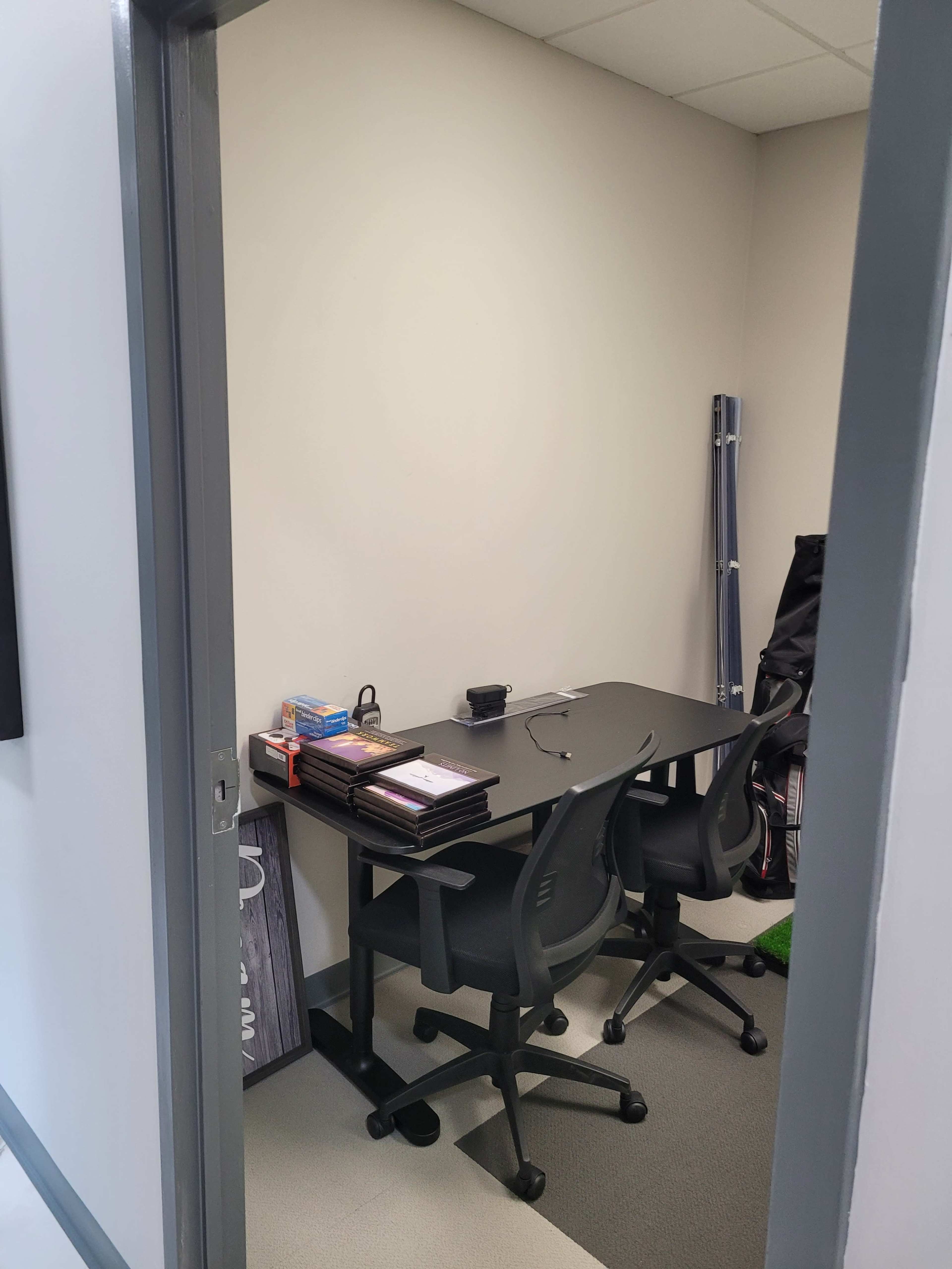 A small office space with a black table and two chairs, along with various items organized on the table and a green area rug on the floor.