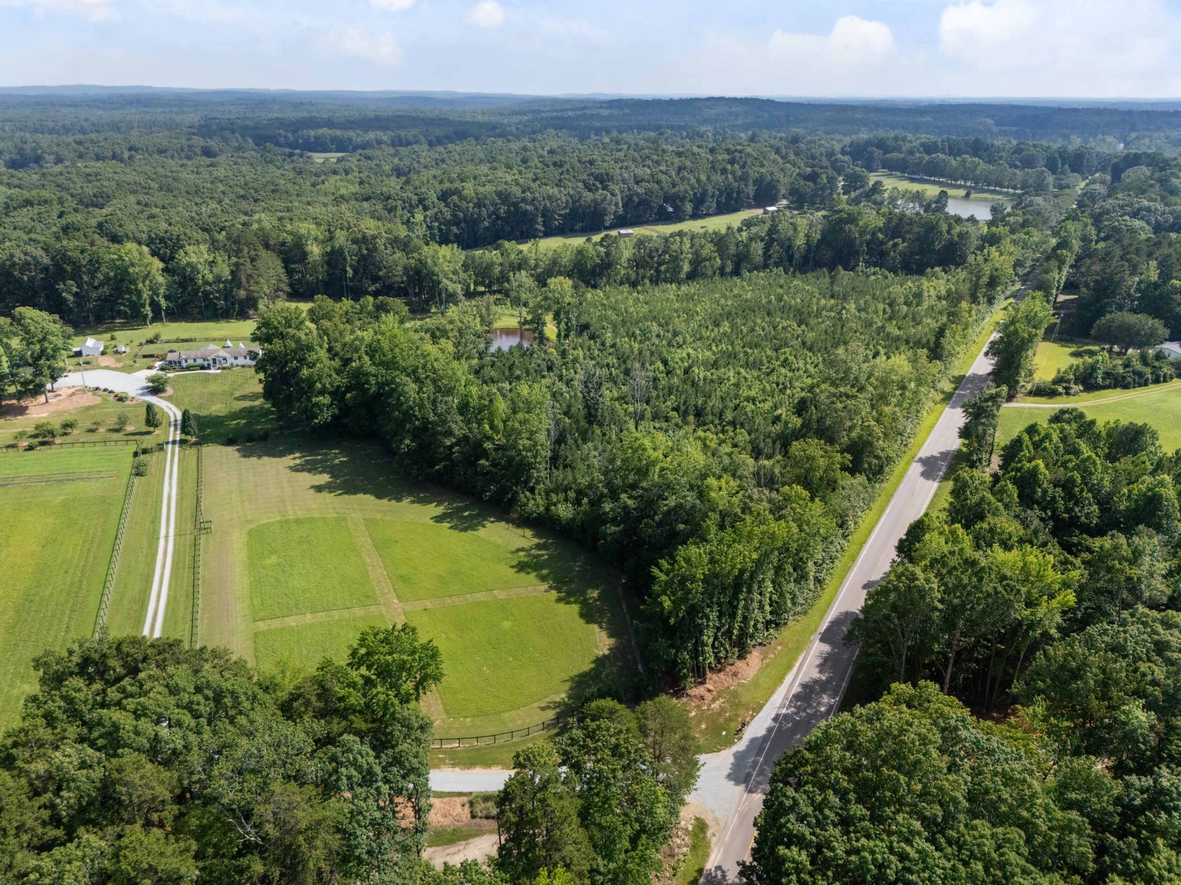 The image shows an aerial view of a lush green landscape featuring a wooded area bordered by a winding road and open fields.
