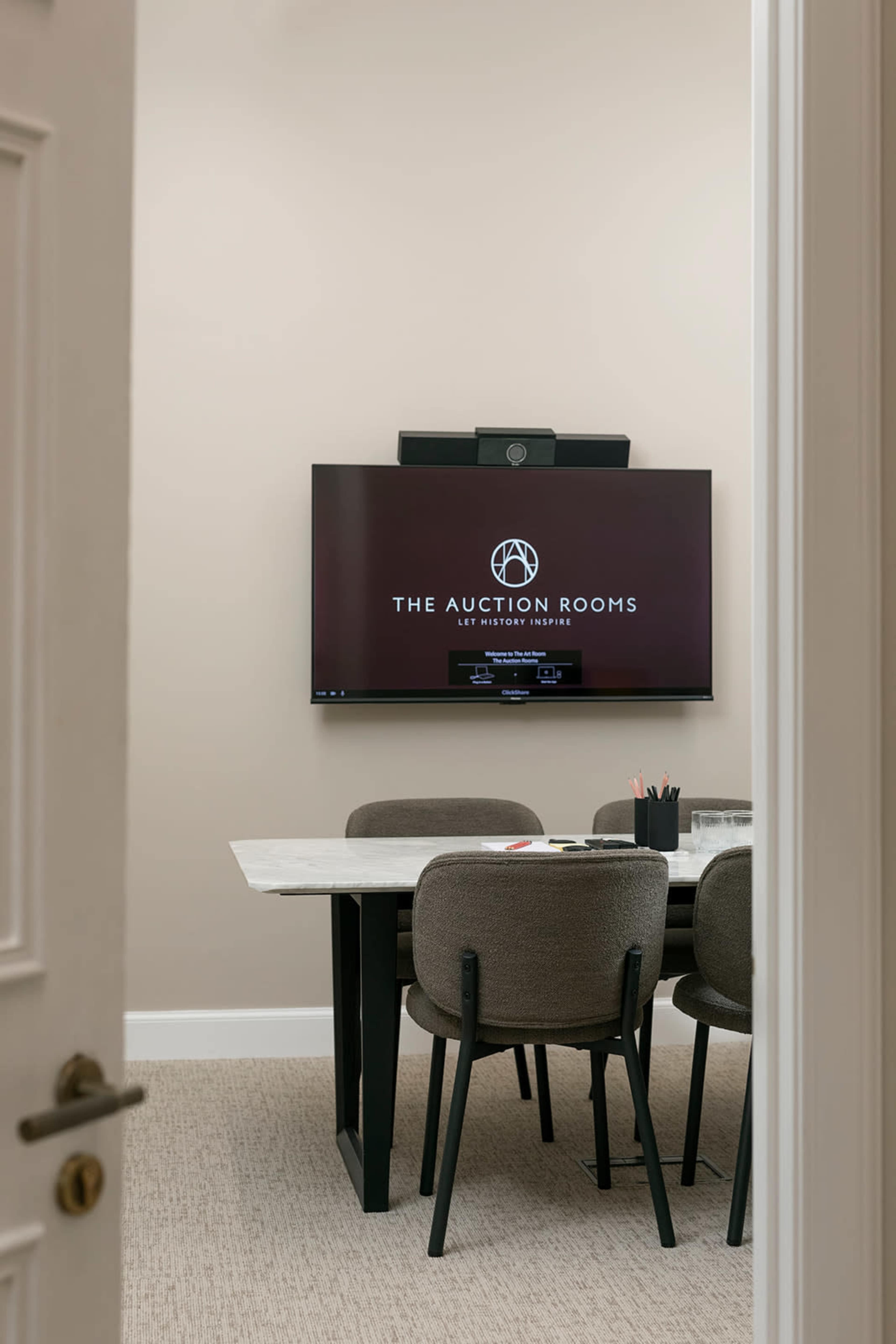 A conference room with a marble table, four gray chairs, and a large television displaying "THE AUCTION ROOMS" on the wall.