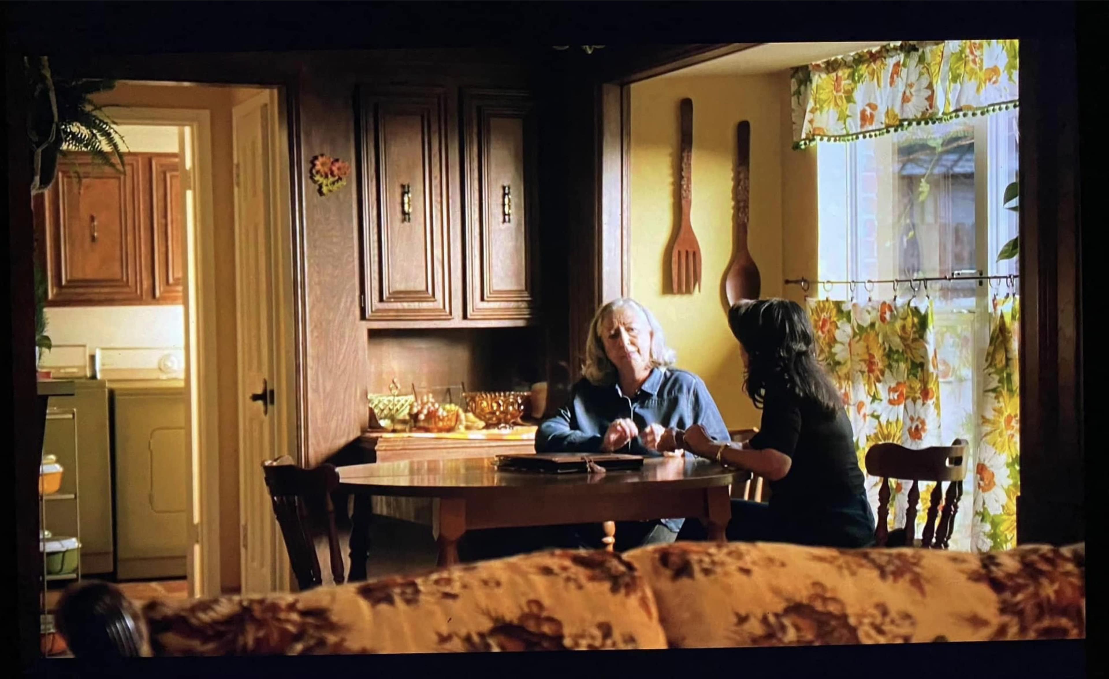 Two women are seated at a table in a warmly lit kitchen, engaged in conversation.