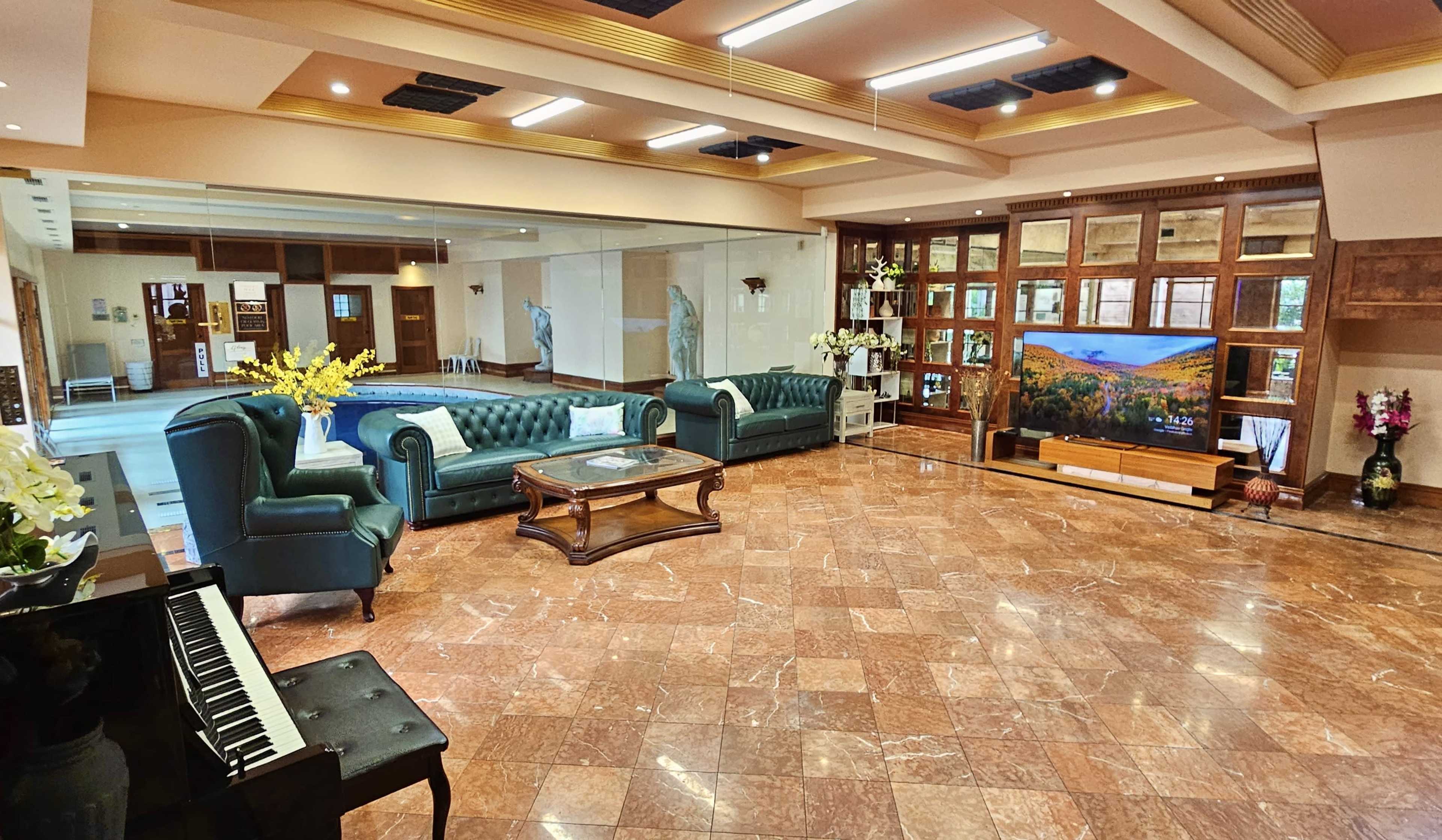 The image shows a spacious and elegantly furnished living room featuring a piano, comfortable couches, a coffee table, and a large television against a wooden wall unit.