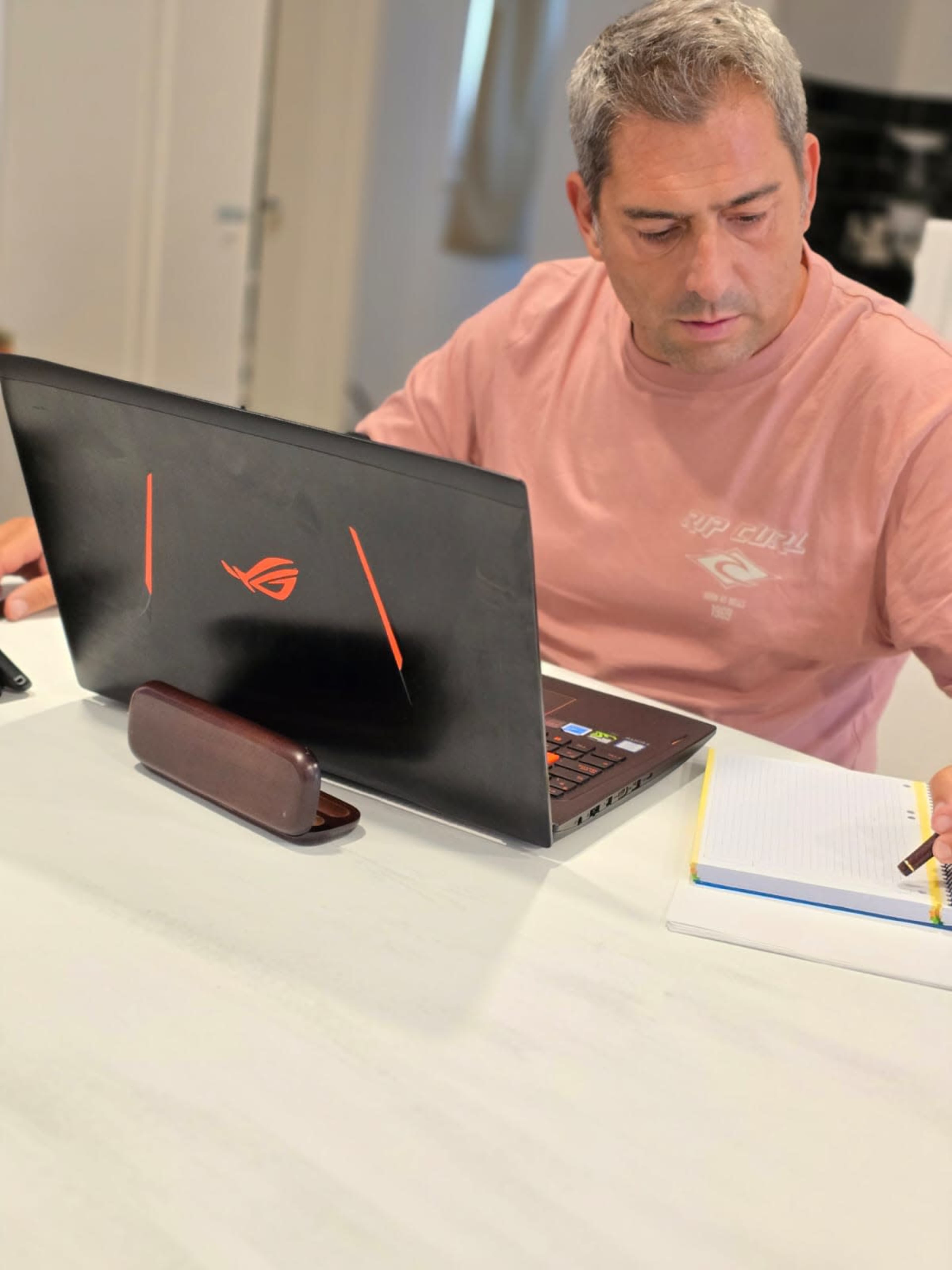 A man sits at a table with a laptop, writing in a notebook.