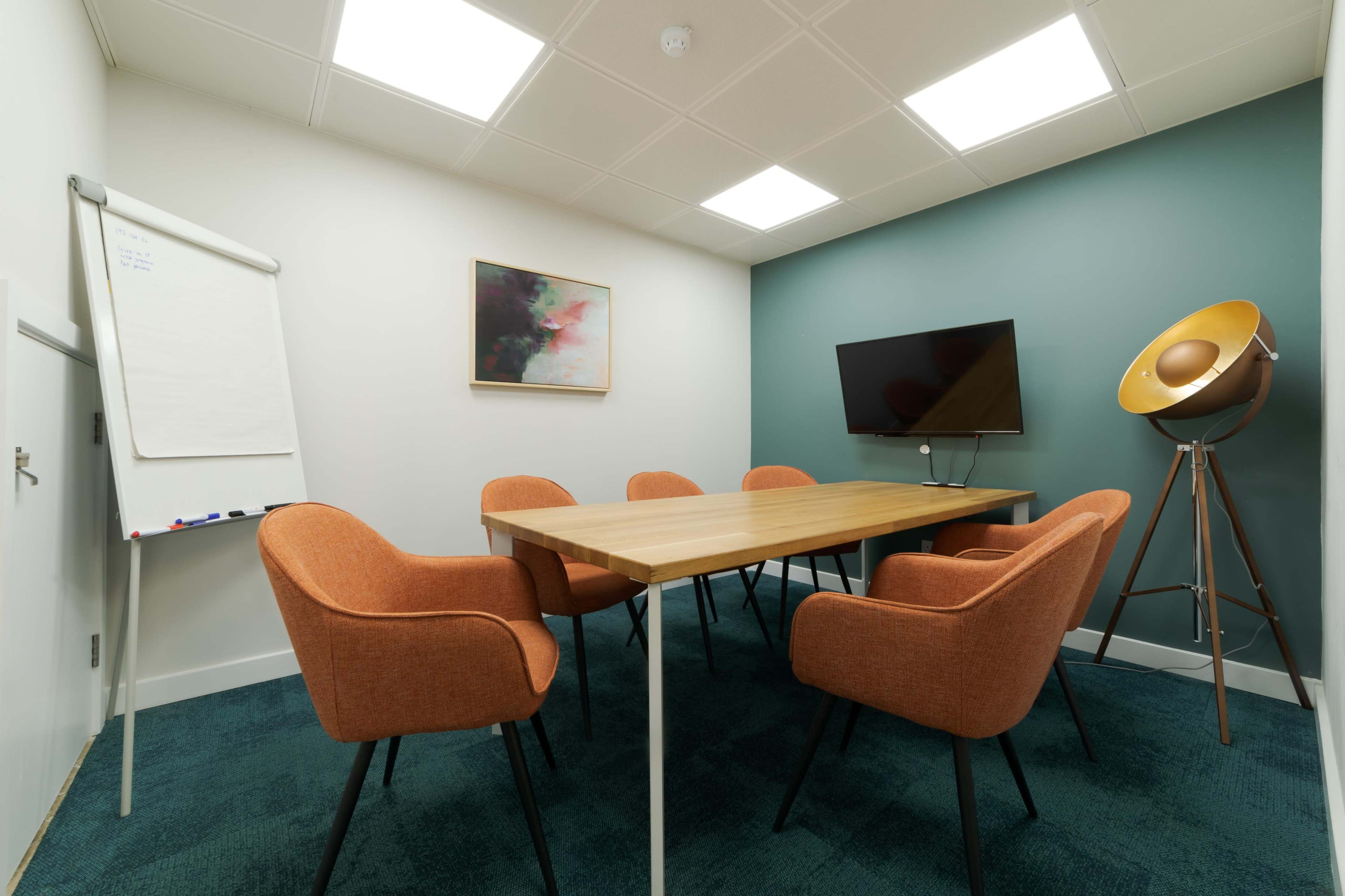 A conference room with a wooden table surrounded by six orange chairs, a whiteboard, a television mounted on the wall, and a decorative floor lamp.