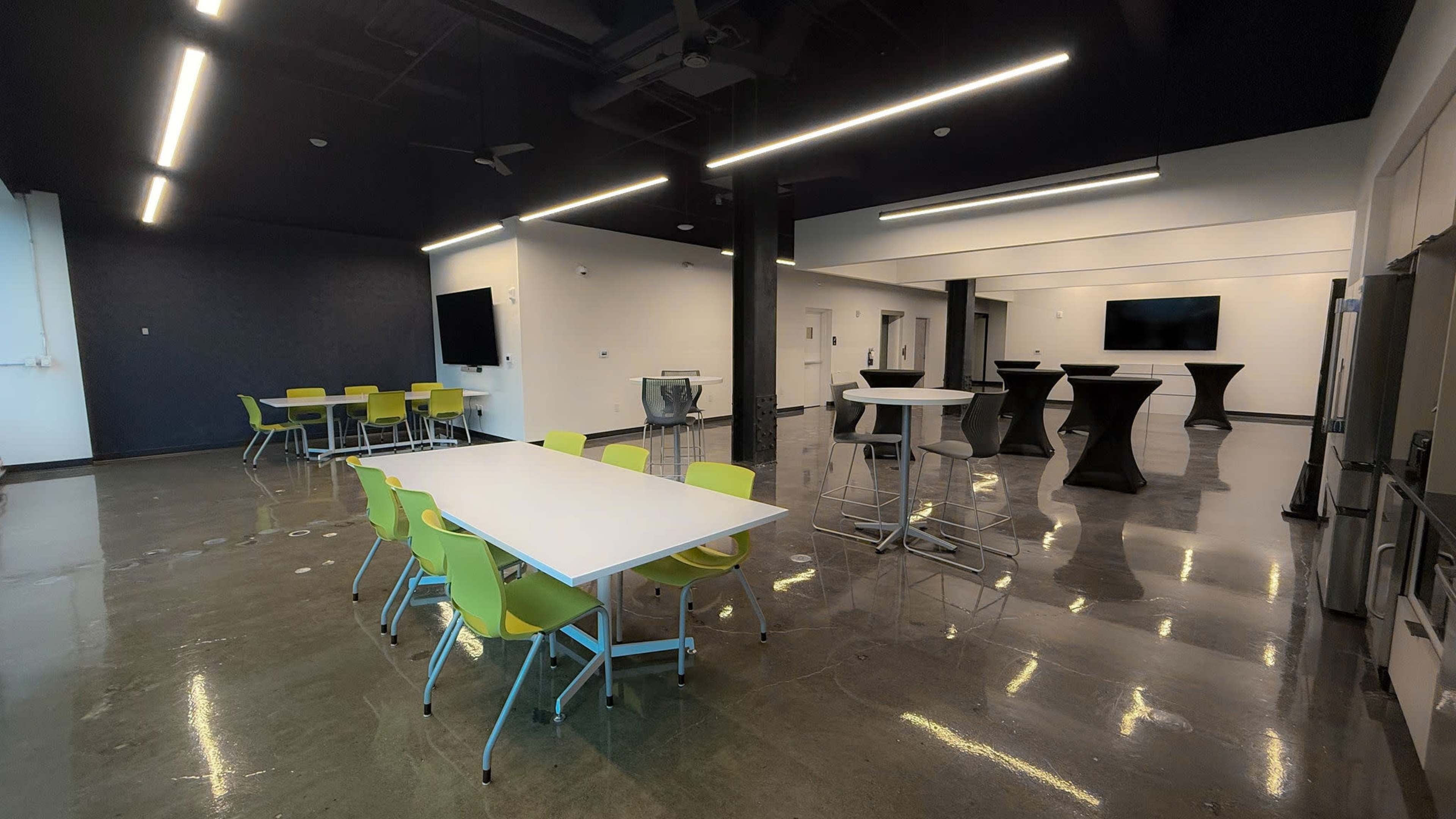 The image shows a modern, well-lit room with a polished concrete floor, featuring various white tables and green chairs arranged for meetings or gatherings.