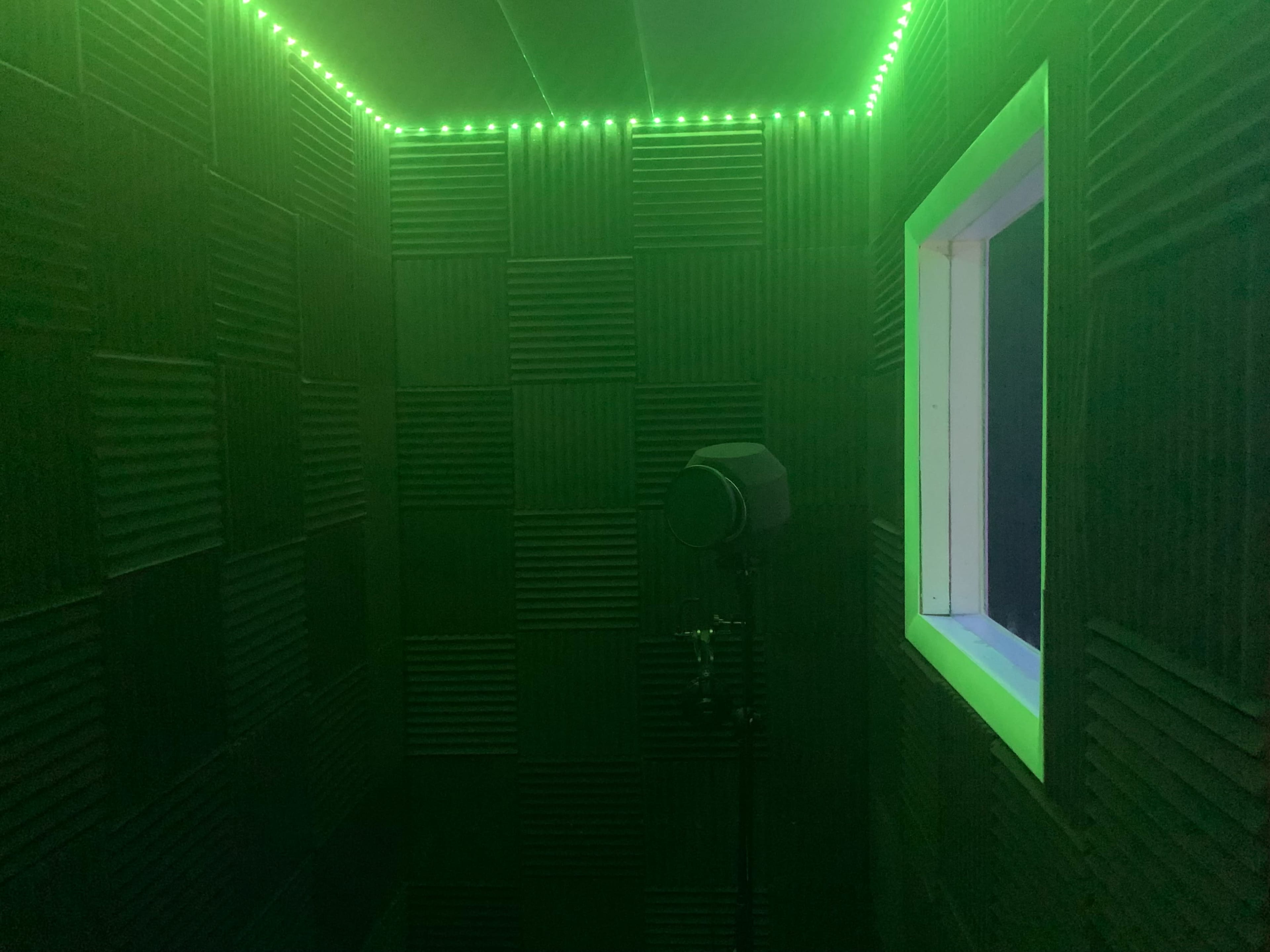 A small room with soundproof walls, illuminated by green LED lights, featuring a microphone stand and a window.