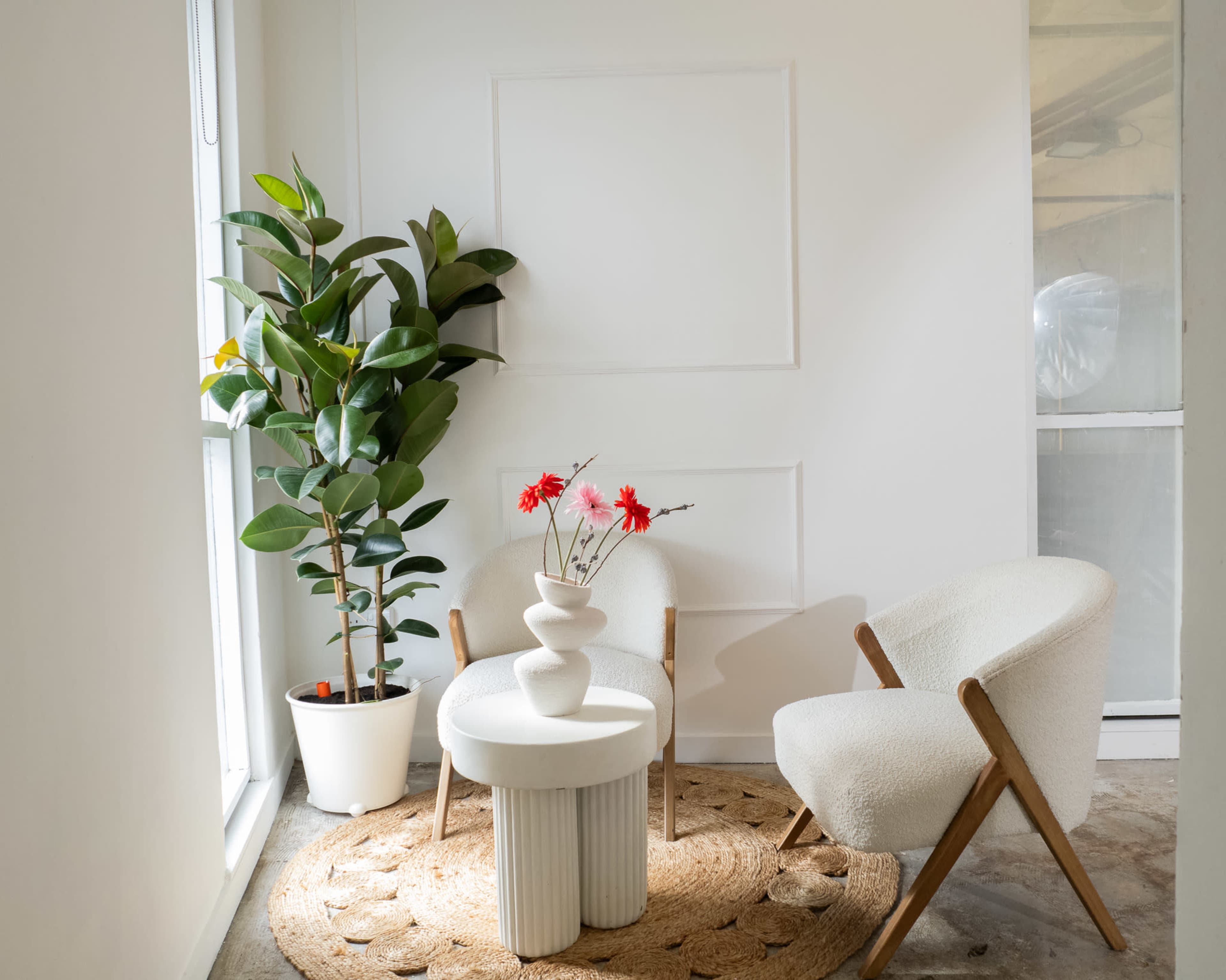 The scene features two white chairs with wooden legs positioned around a round table, accompanied by a potted plant and a vase with flowers, all set in a well-lit room with white walls.